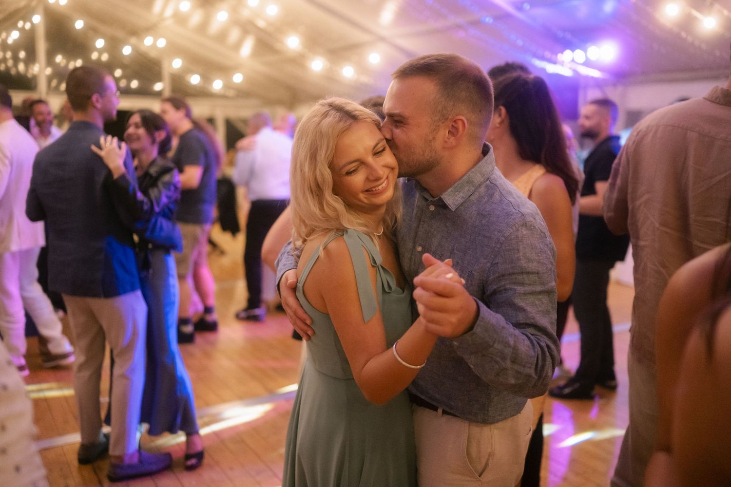 Couples dancing together at an indoor wedding reception with purple lighting and string lights overhead.