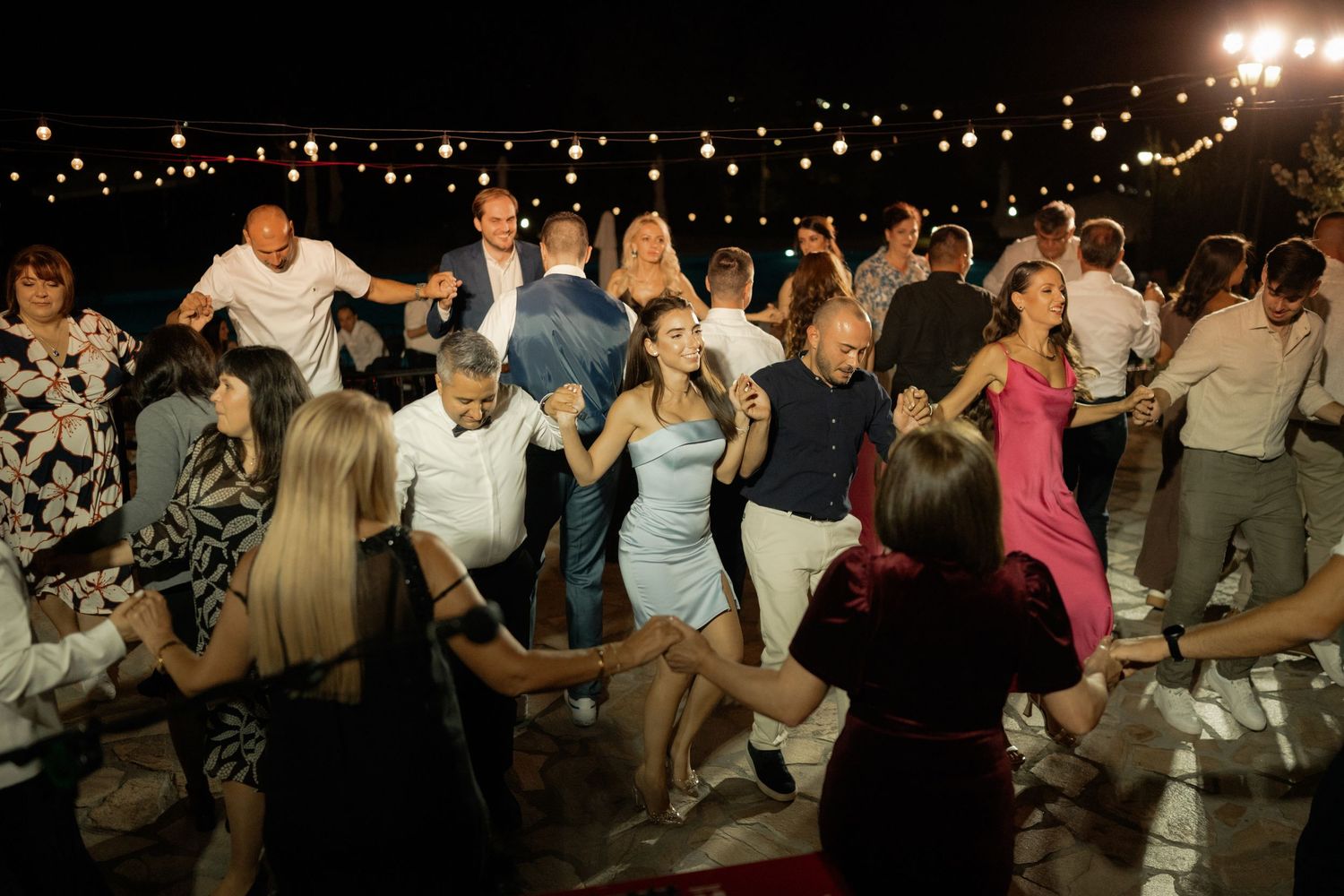 Wedding guests dance energetically on an outdoor dance floor at night with twinkling lights in the background.