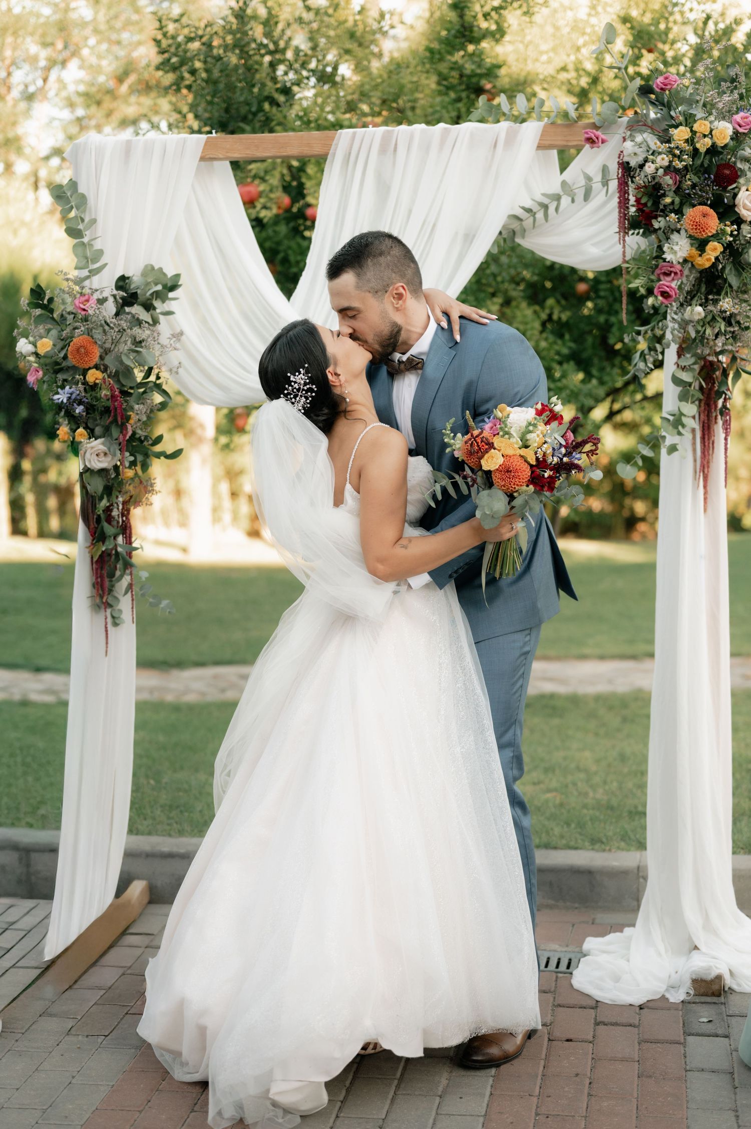 Romantic wedding couple shares intimate moments under white draped pergola decorated with colorful autumn florals.