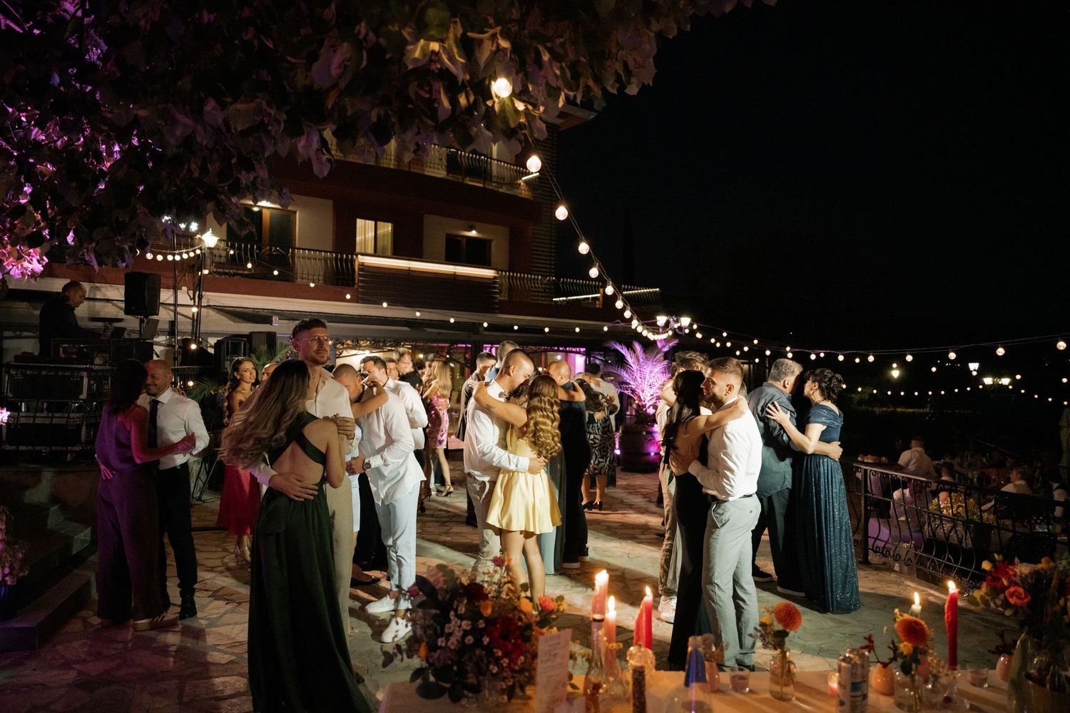 People dance and celebrate at an outdoor nighttime wedding reception with string lights overhead and glowing decorations.