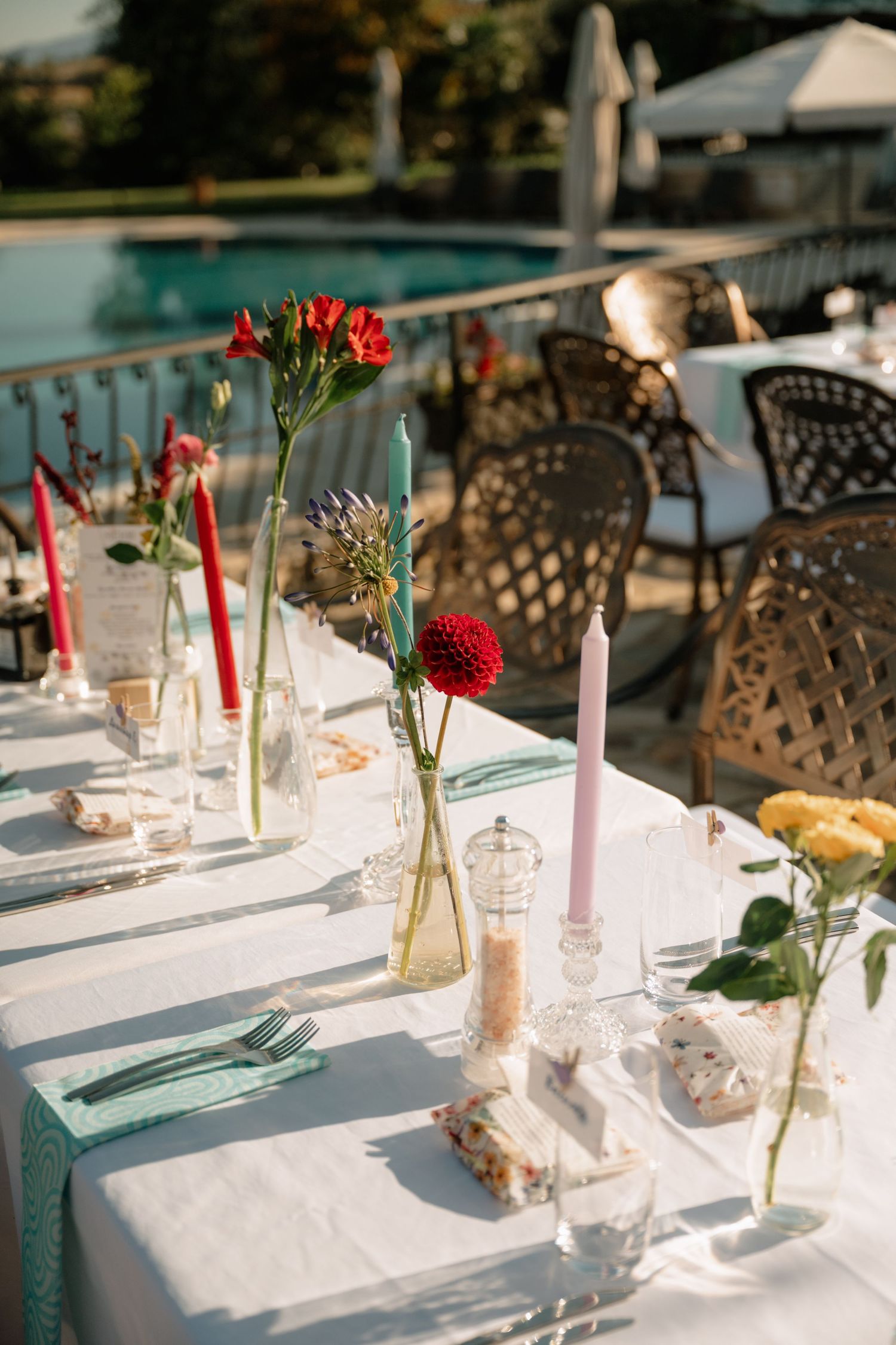 Elegant outdoor table settings with white linens, glass bottles, and red floral centerpieces in golden sunlight.