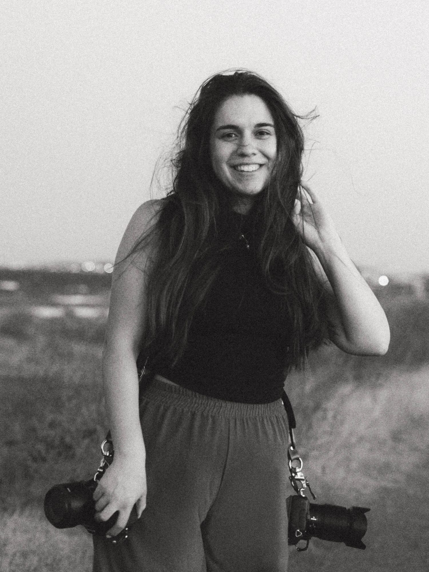 Smiling woman with long hair holding two cameras outdoors in a black and white portrait photo.