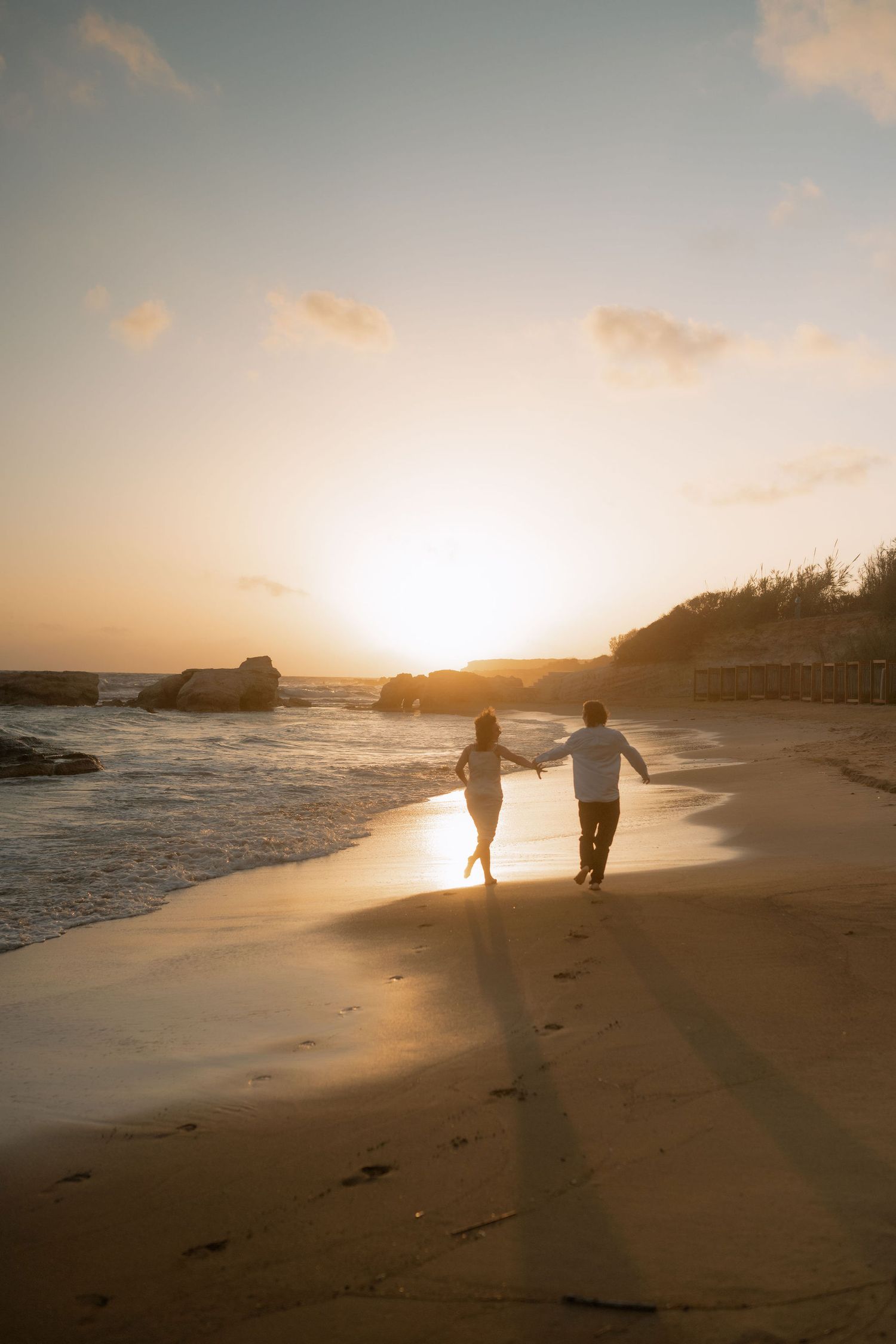 Couple holding hands walking on sandy beach at golden sunset with ocean waves and rocky coastline in background.
