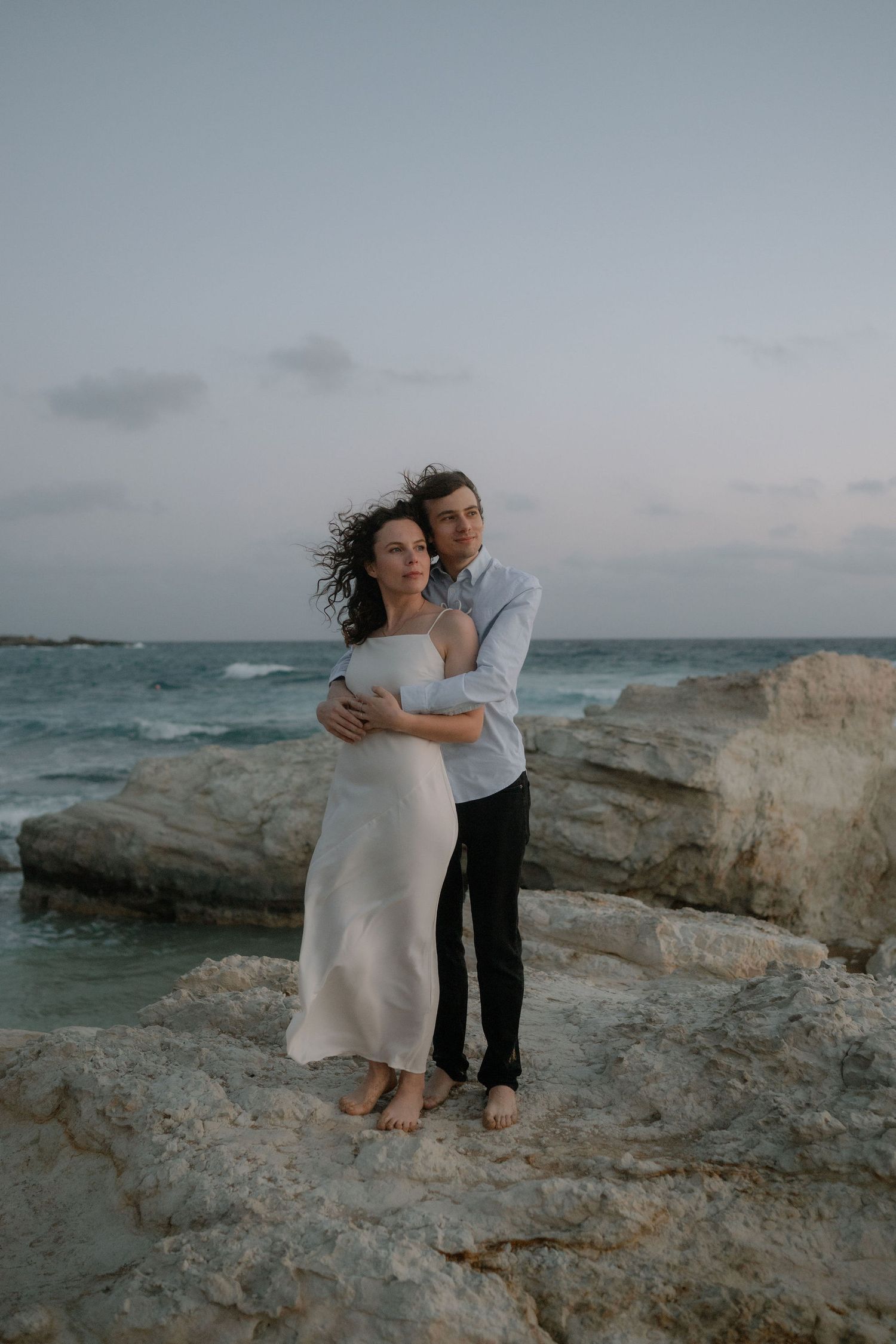 Couple embracing on rocky seaside at dusk, woman in white dress, man in blue shirt, with turquoise ocean waves behind them.