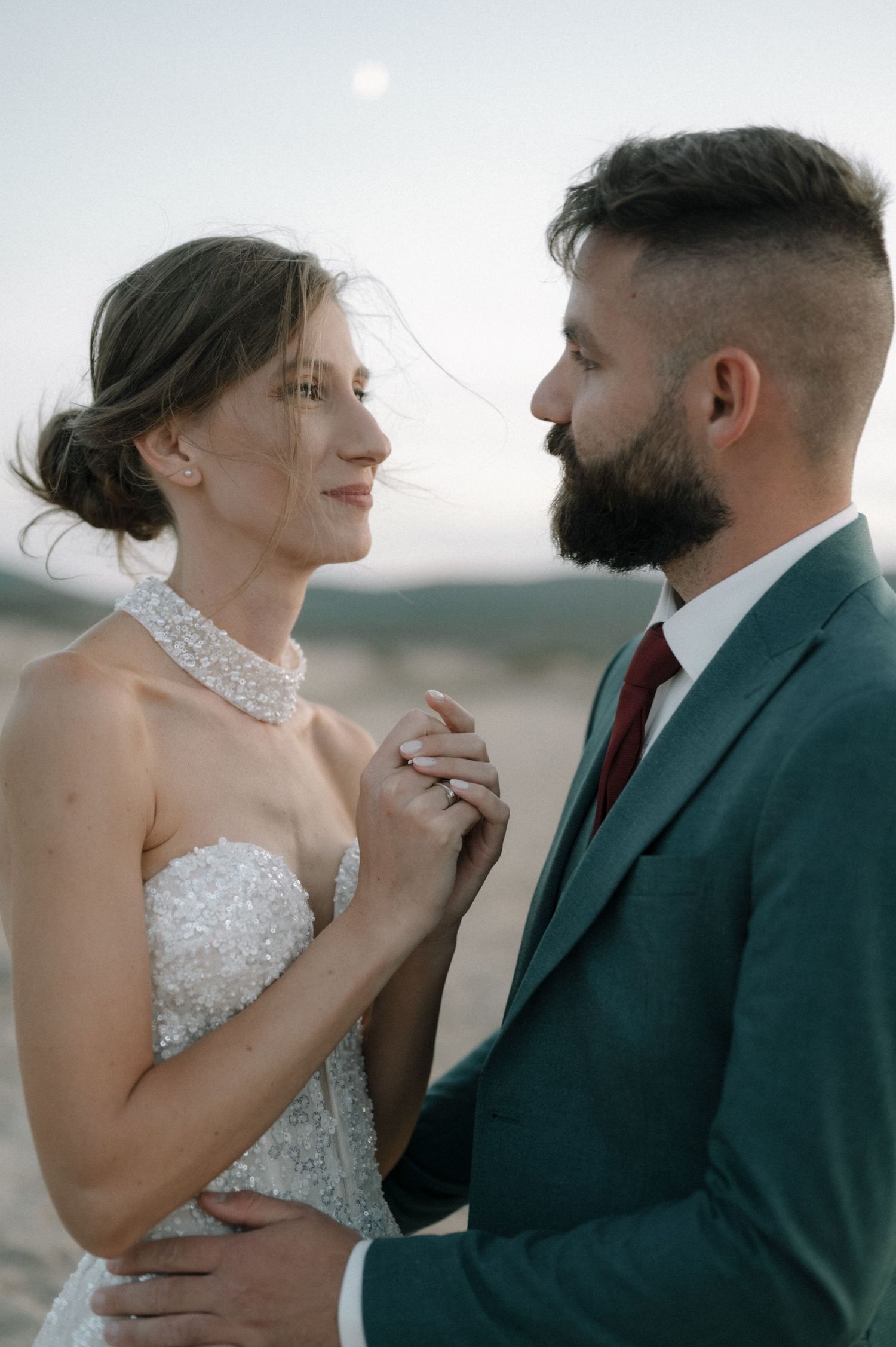 A bride and groom share a tender moment holding hands outdoors at dusk, she in a beaded gown, he in a teal suit.