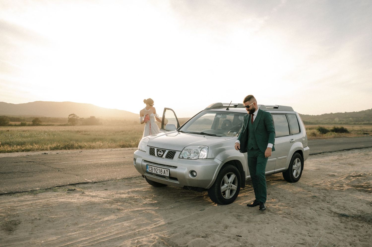 Bride and groom posing beside a silver Nissan X-Trail SUV on a dirt road at golden sunset.