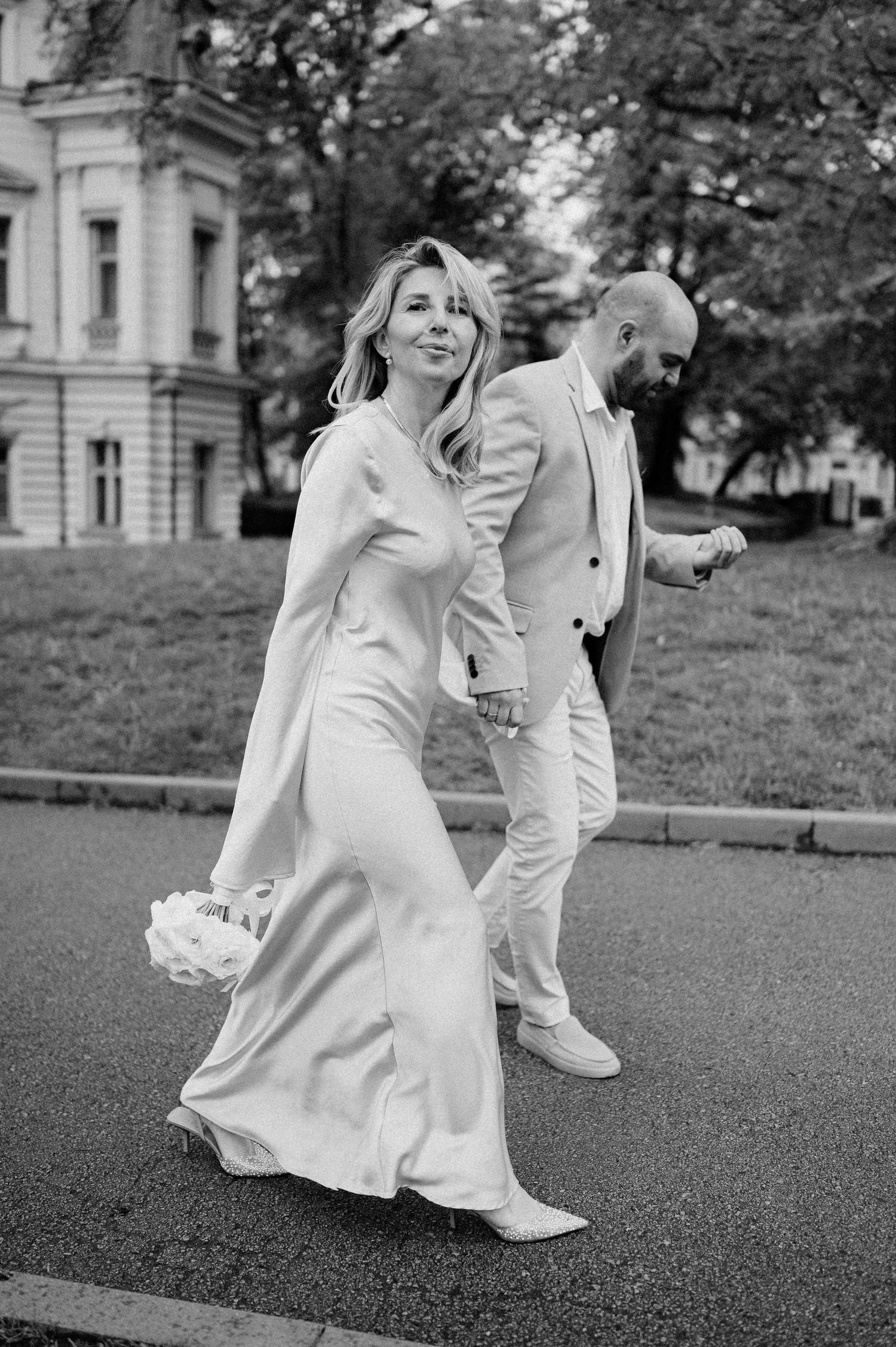 A couple in white attire dances playfully on a paved path.