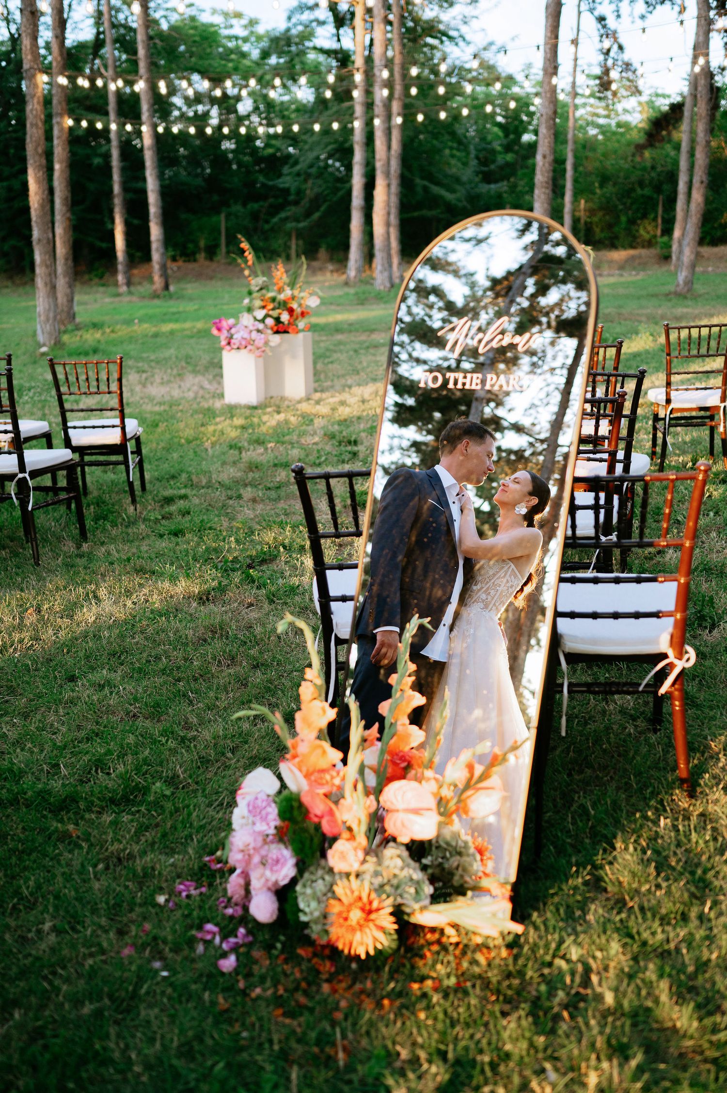 Outdoor wedding ceremony setup with wooden chairs and colorful floral arrangements in a wooded setting at sunset.