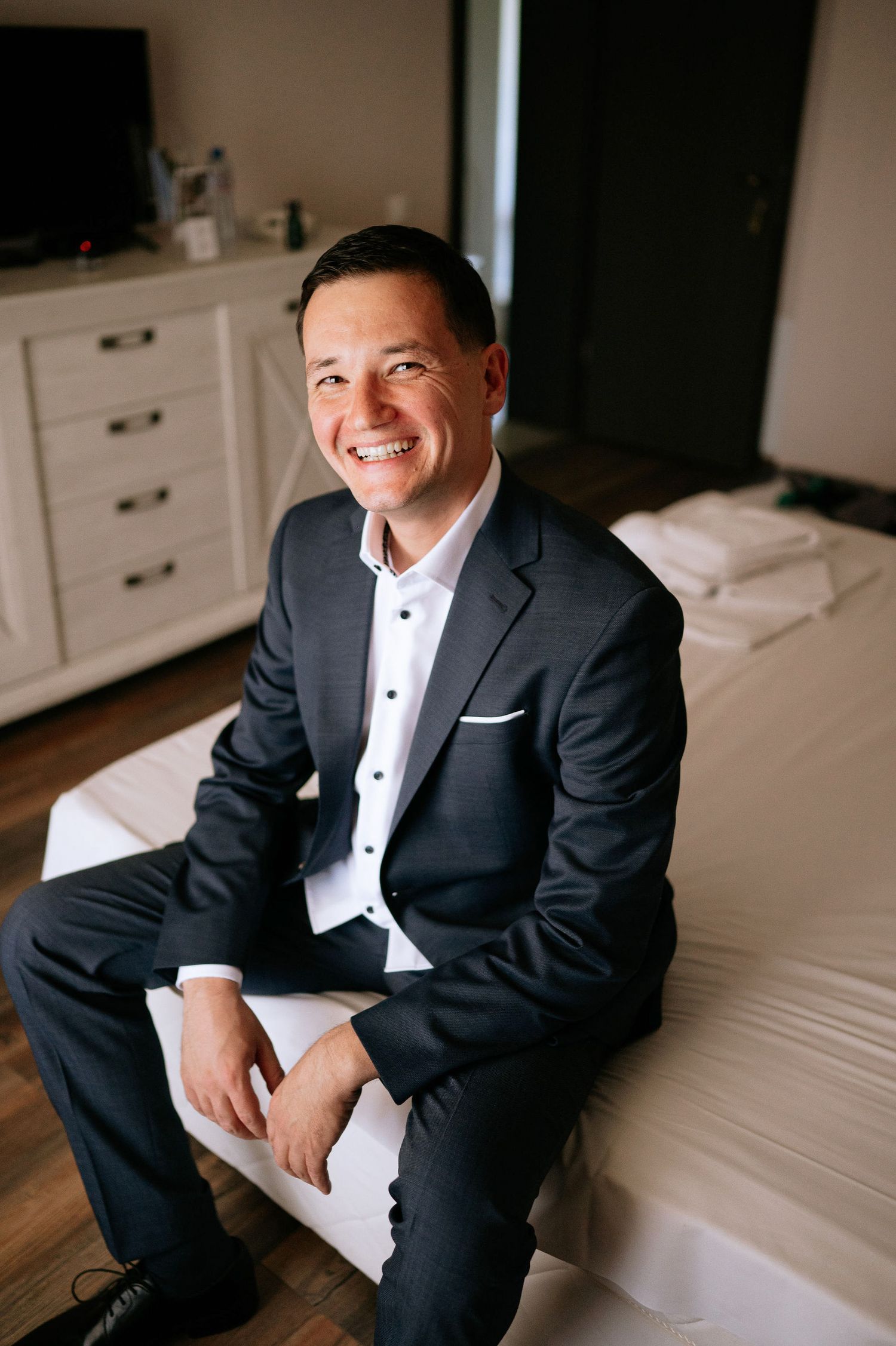 Person in formal attire sitting on white bedding with a genuine smile.