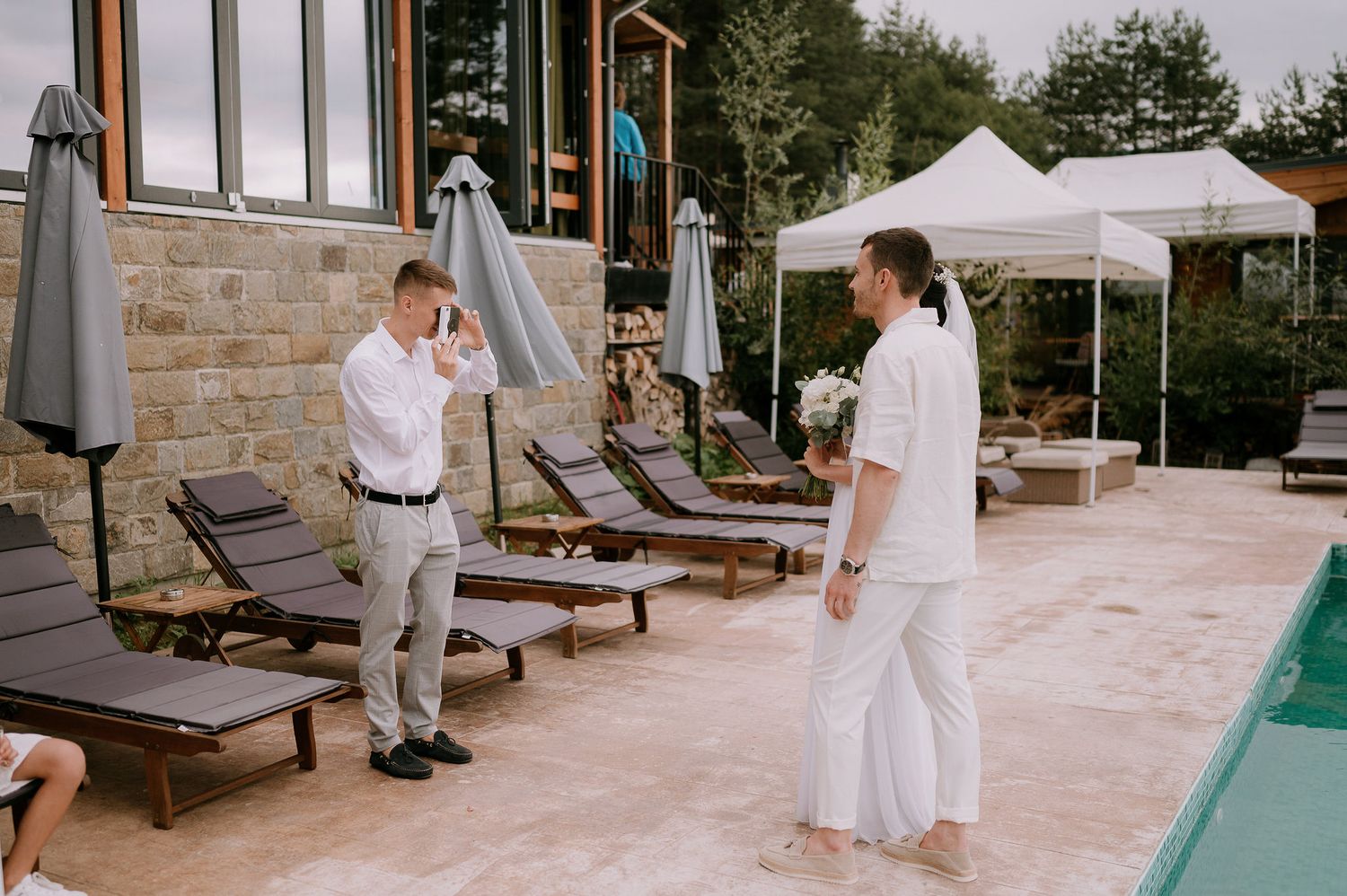 A poolside wedding venue with white lounge chairs and tents set up on a stone patio.