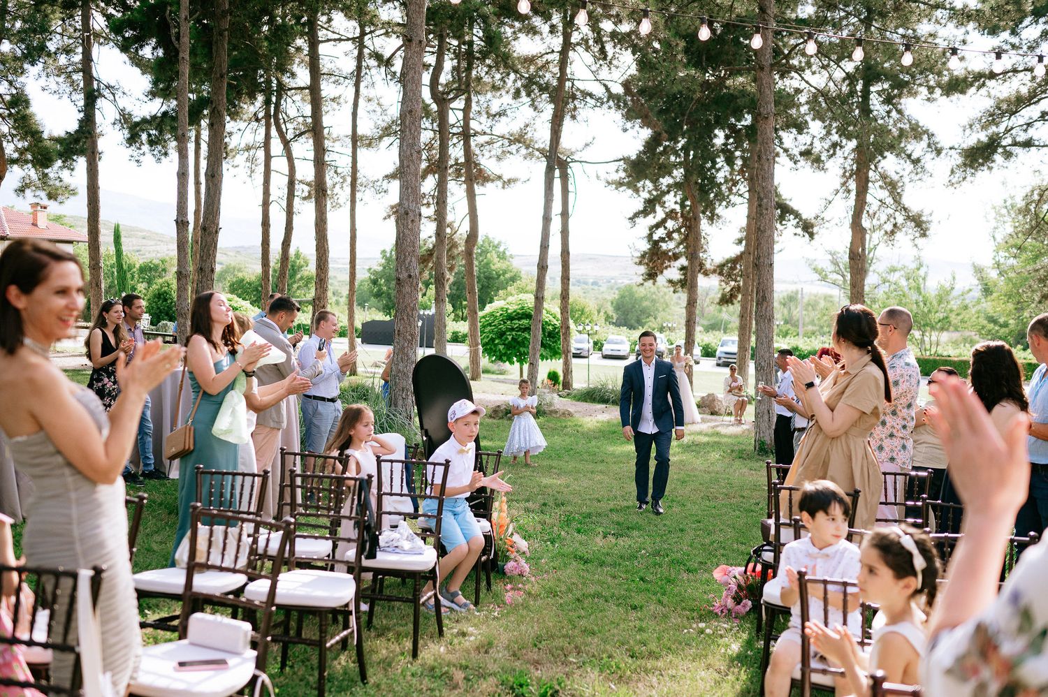 Wedding guests seated on wooden chairs during outdoor ceremony in wooded setting.