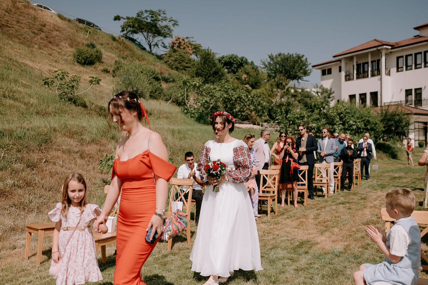 Wedding ceremony procession walking down an outdoor aisle with guests watching from wooden chairs.