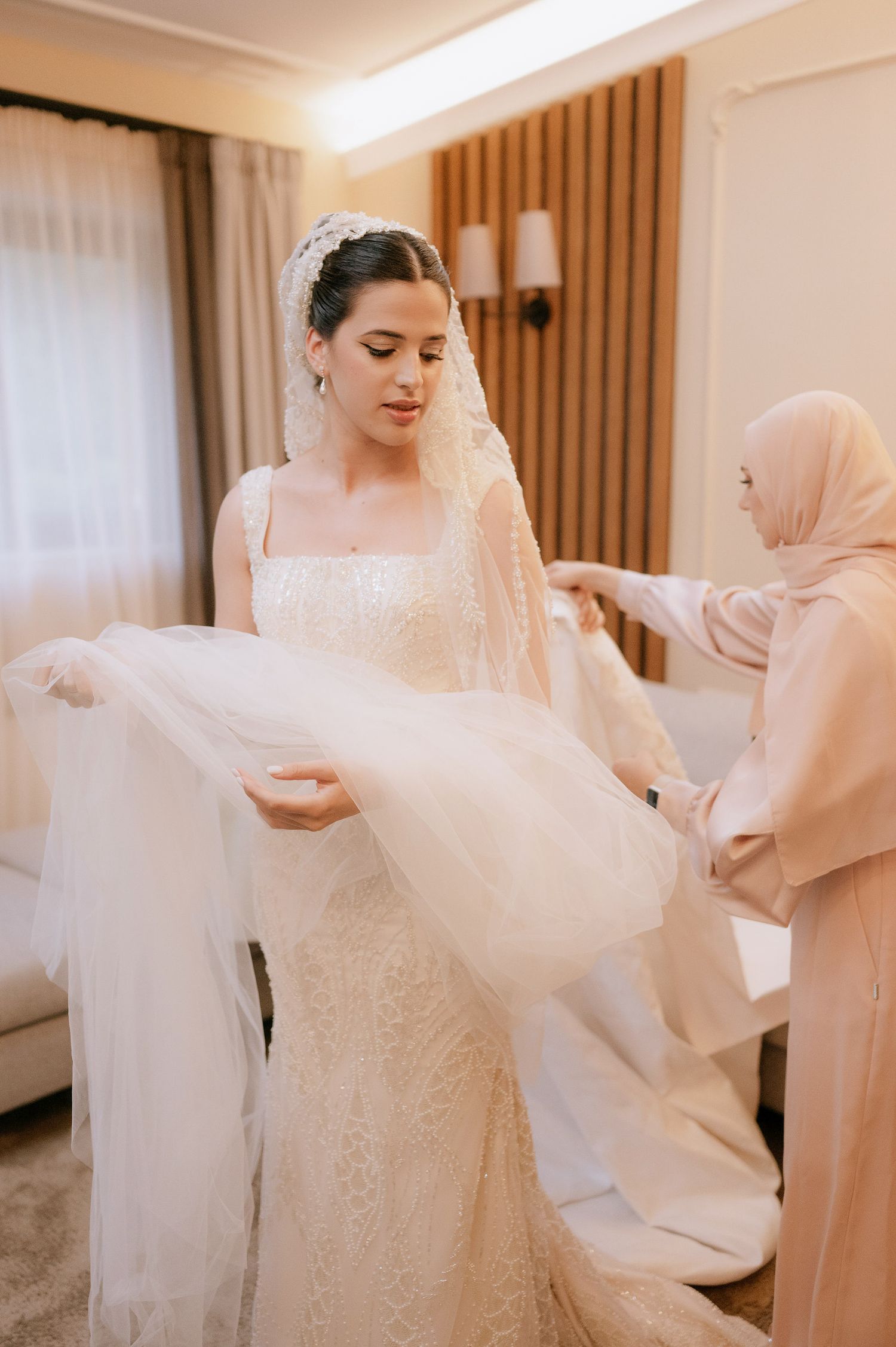 A bride in a white lace gown gets help with her wedding veil from attendants in a warmly lit dressing room.
