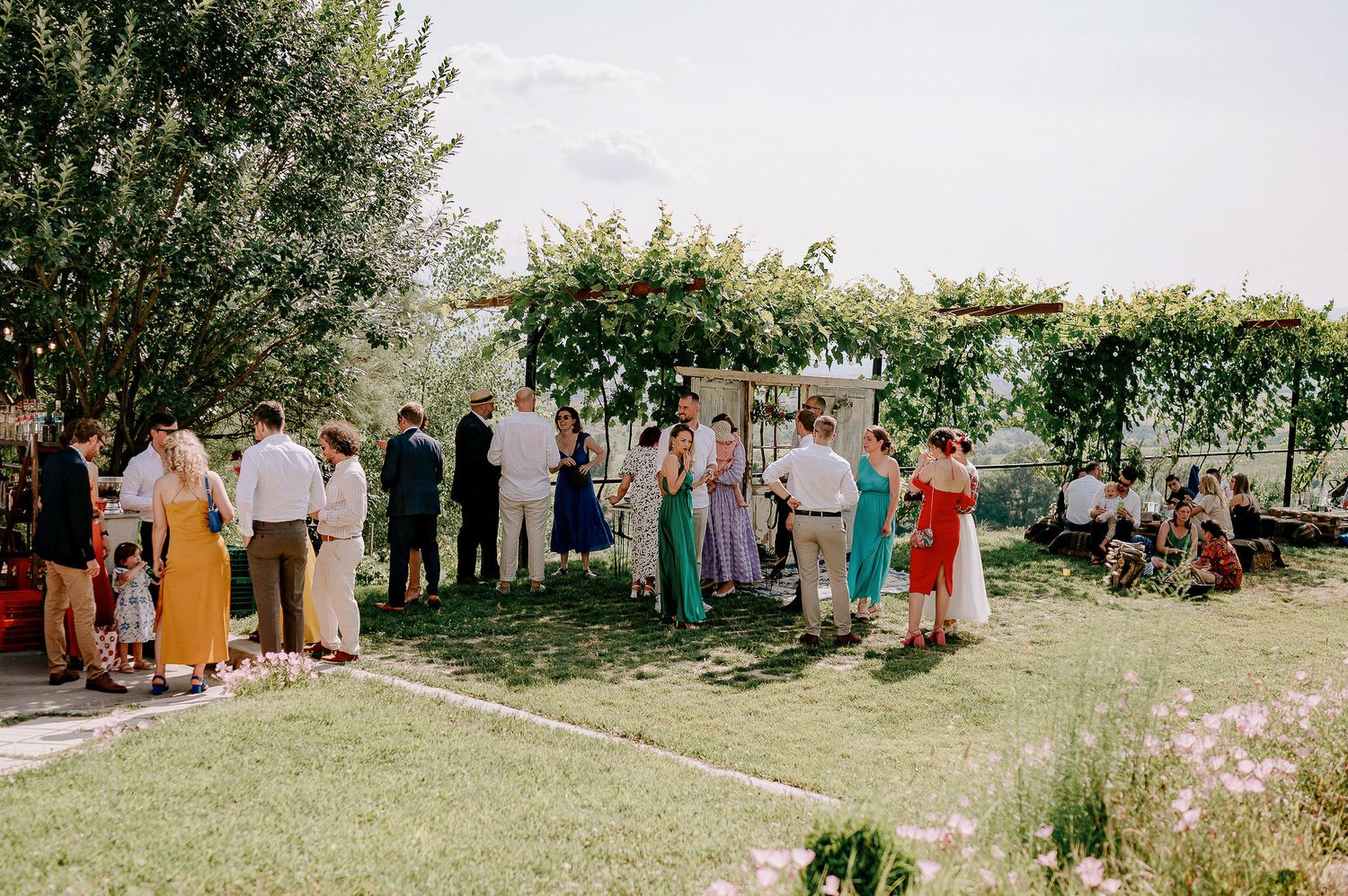 Outdoor wedding reception with guests gathered on a grassy lawn surrounded by trees.