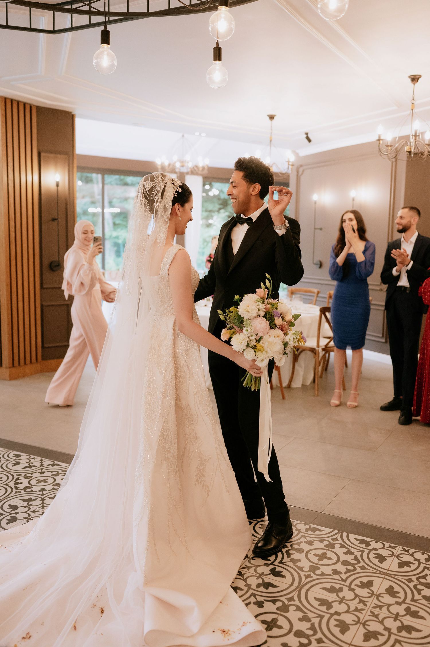 Wedding guests celebrate in a bright reception hall with decorative flooring and modern lighting fixtures.