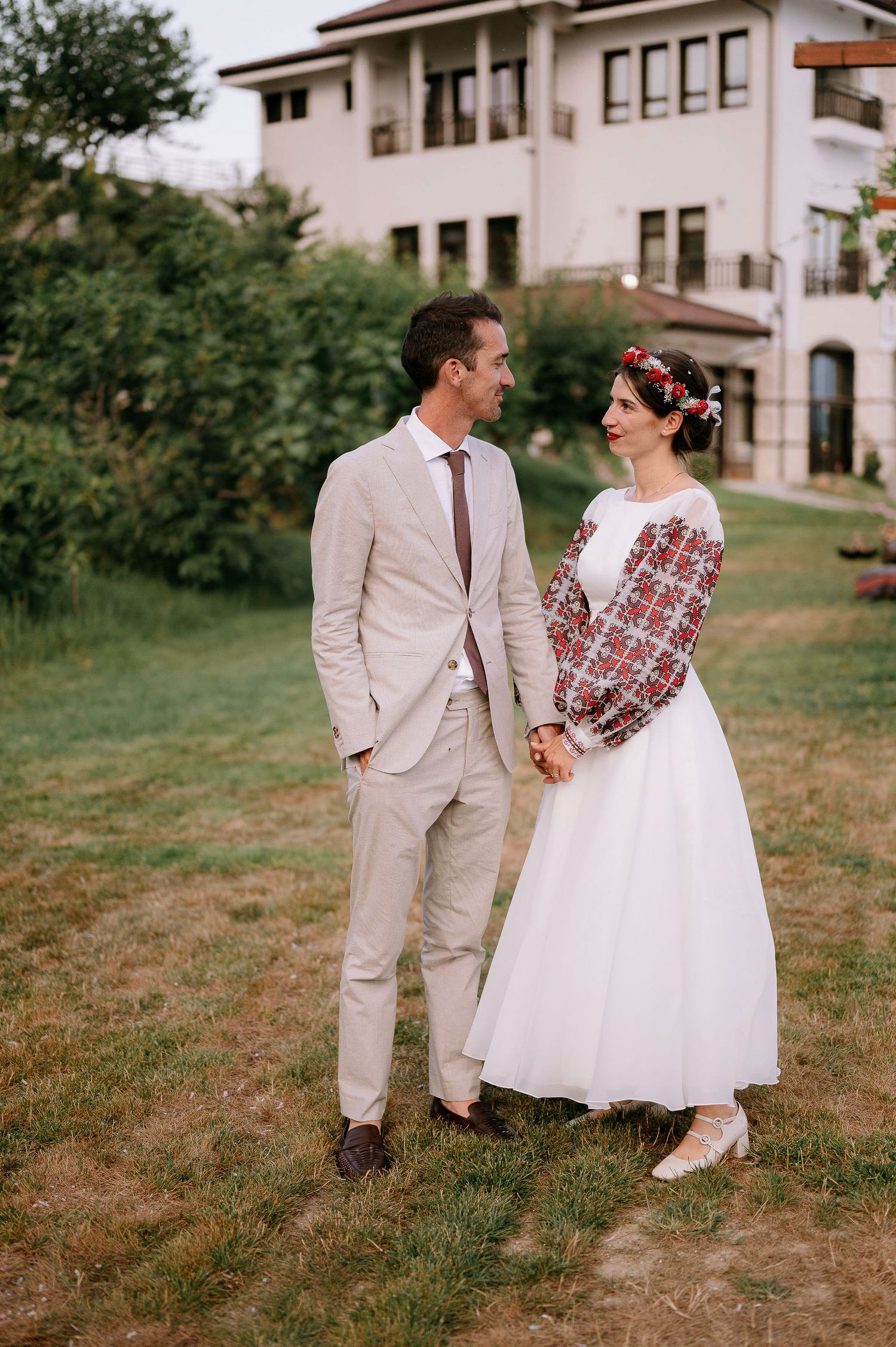 A couple in vintage wedding attire poses together on a grassy slope near a white building.