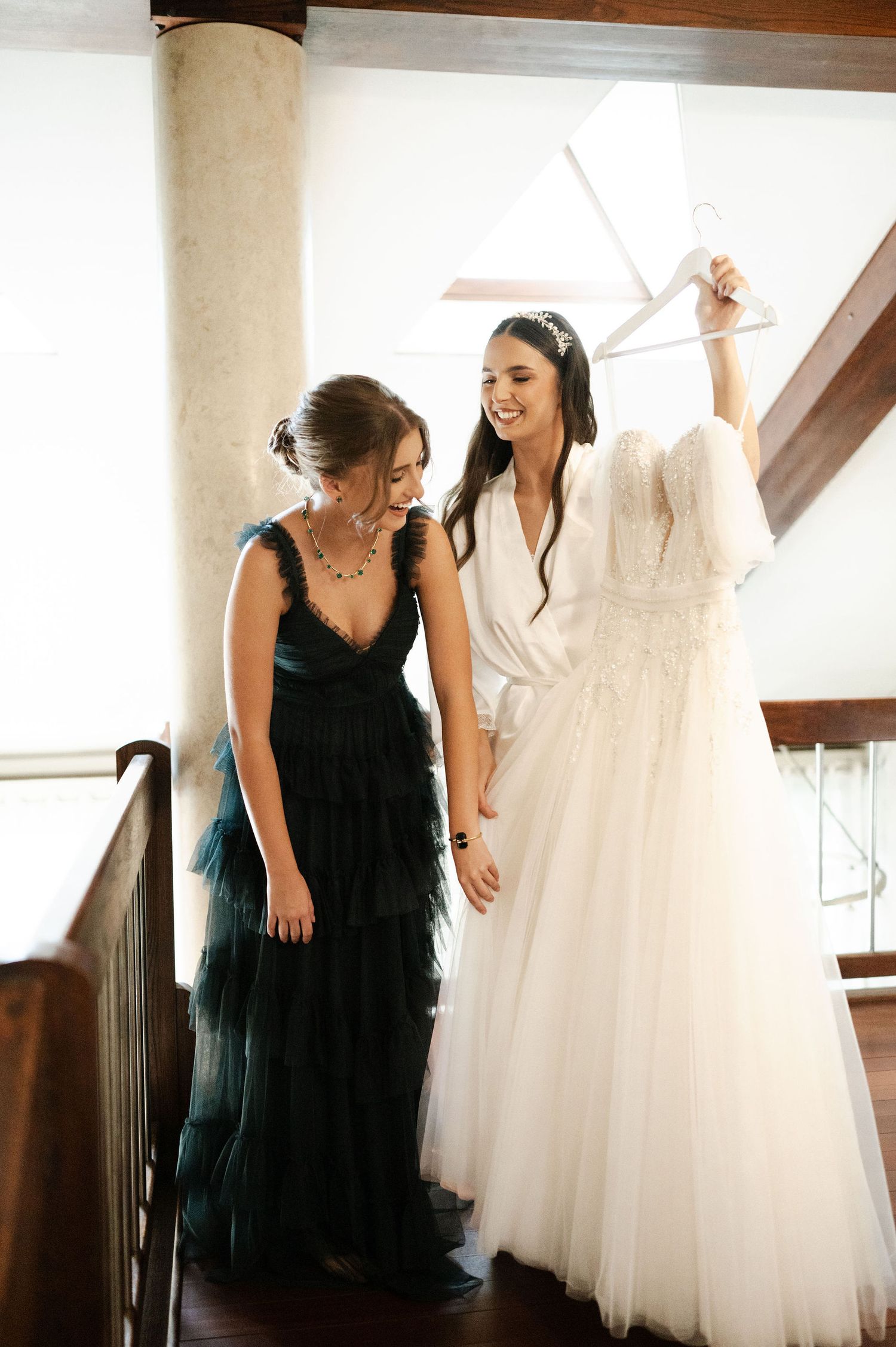 Two women admire a white wedding dress hanging from above.