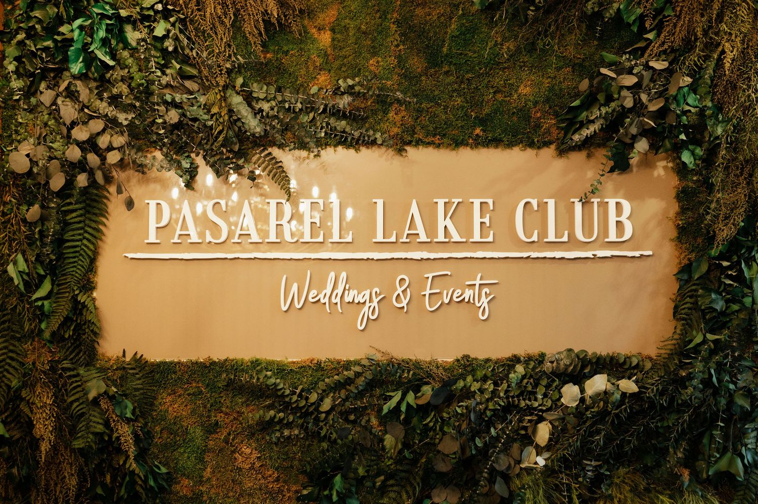 Rustic outdoor venue sign surrounded by lush greenery and ambient lighting.