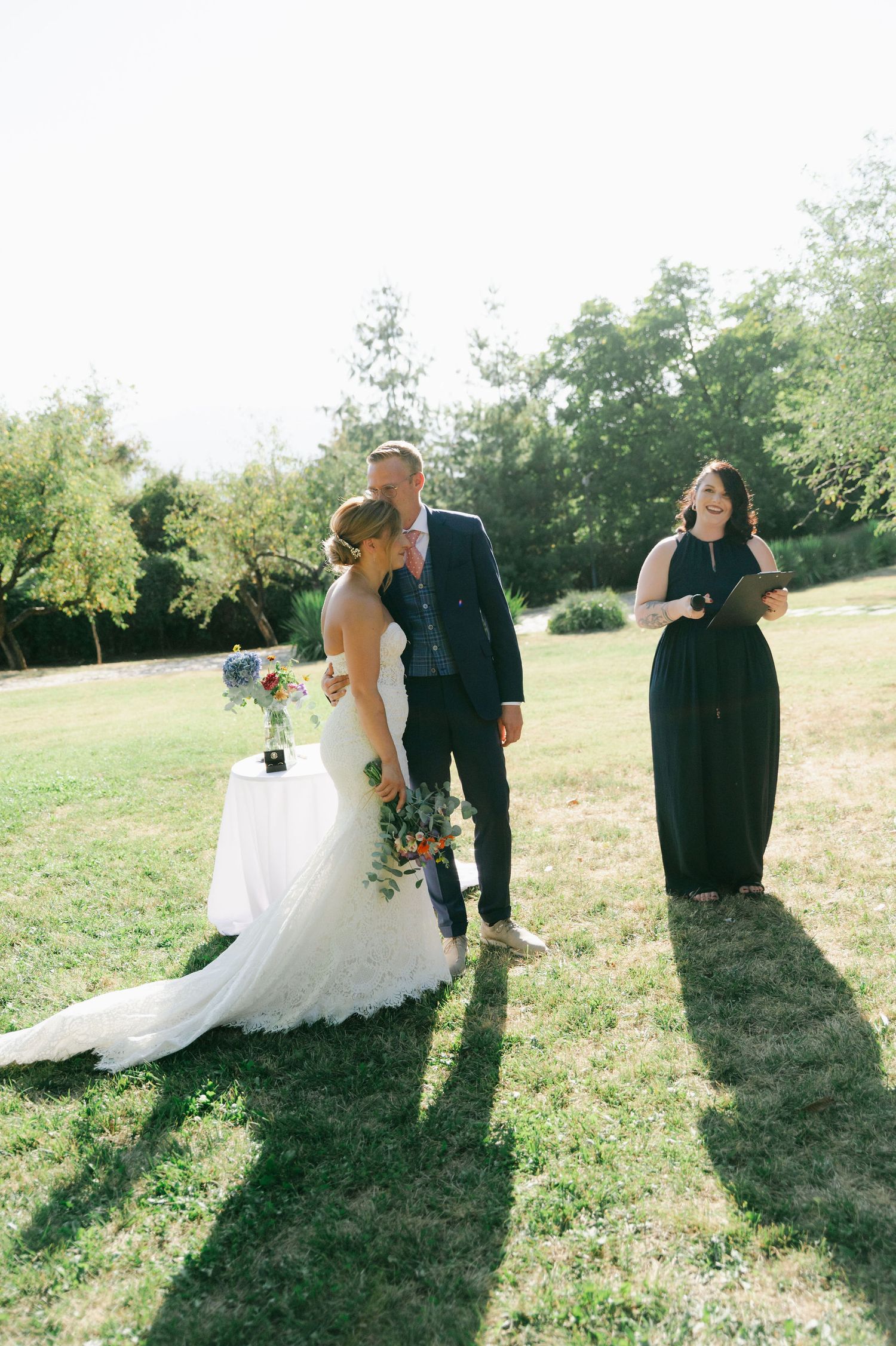 Wedding ceremony participants interact in a sunlit field surrounded by trees.