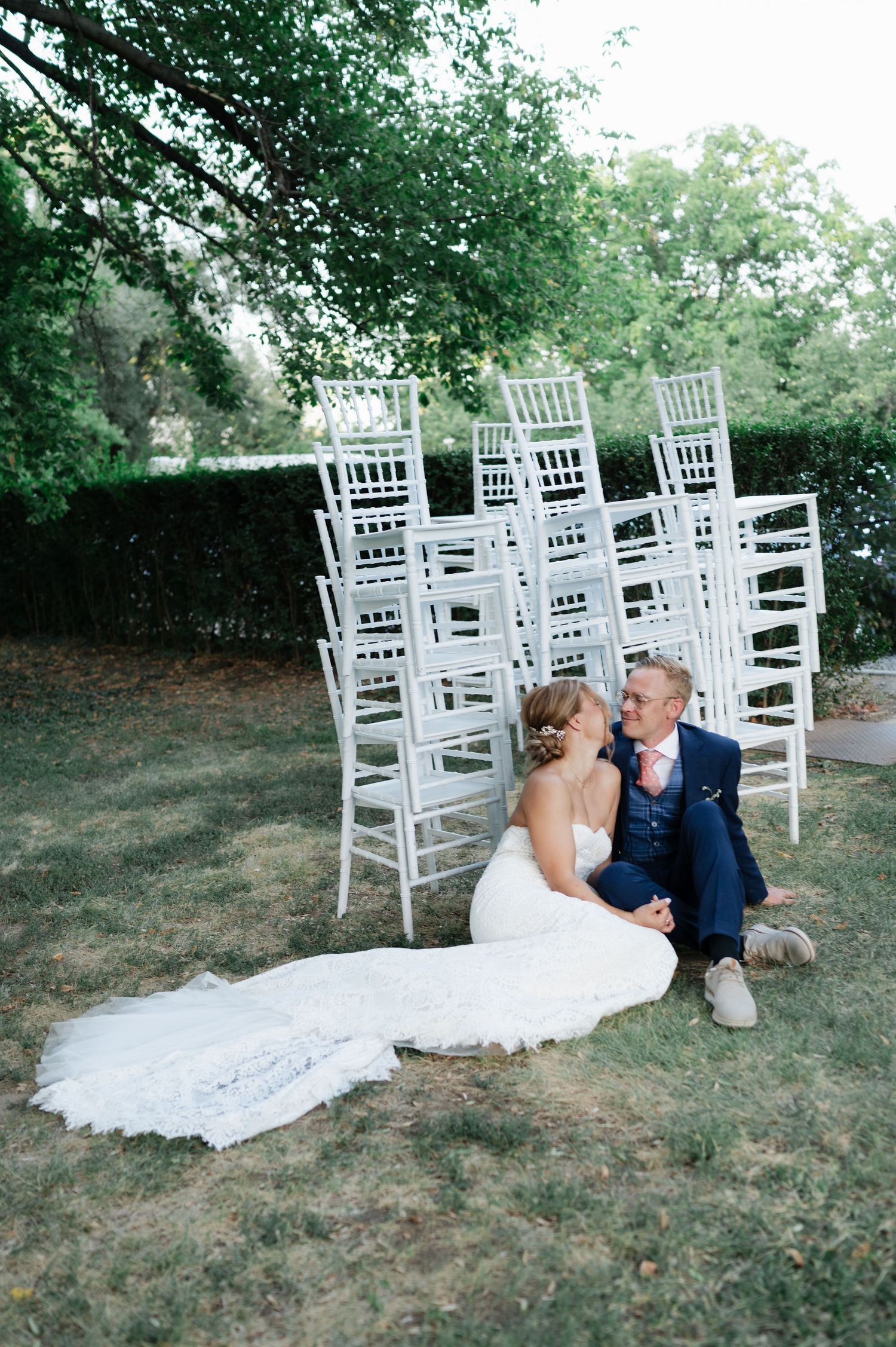 Newlyweds share tender moments on lawn near white ceremony chairs during outdoor photo session.