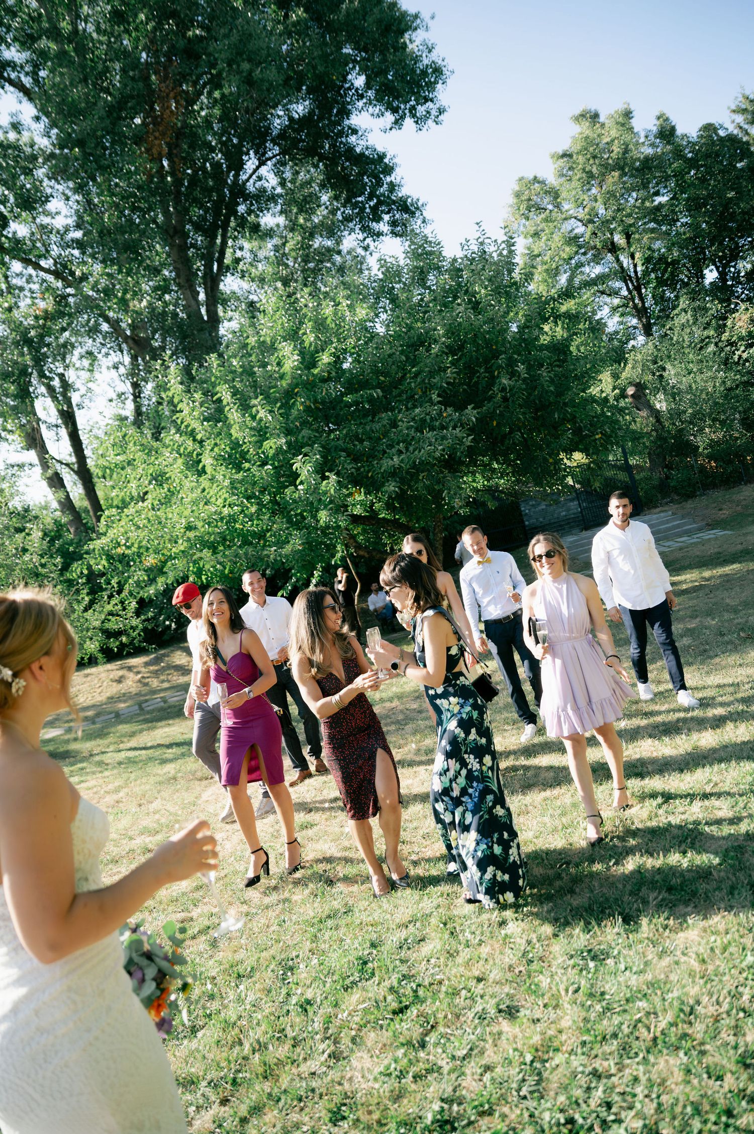 Wedding party members stand in a line on a grassy hillside during a sunny outdoor celebration.