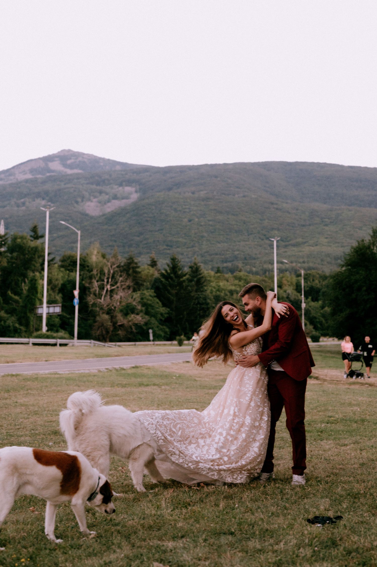 Couple dancing in a field with mountains and dogs, wearing burgundy suit and blush lace gown.