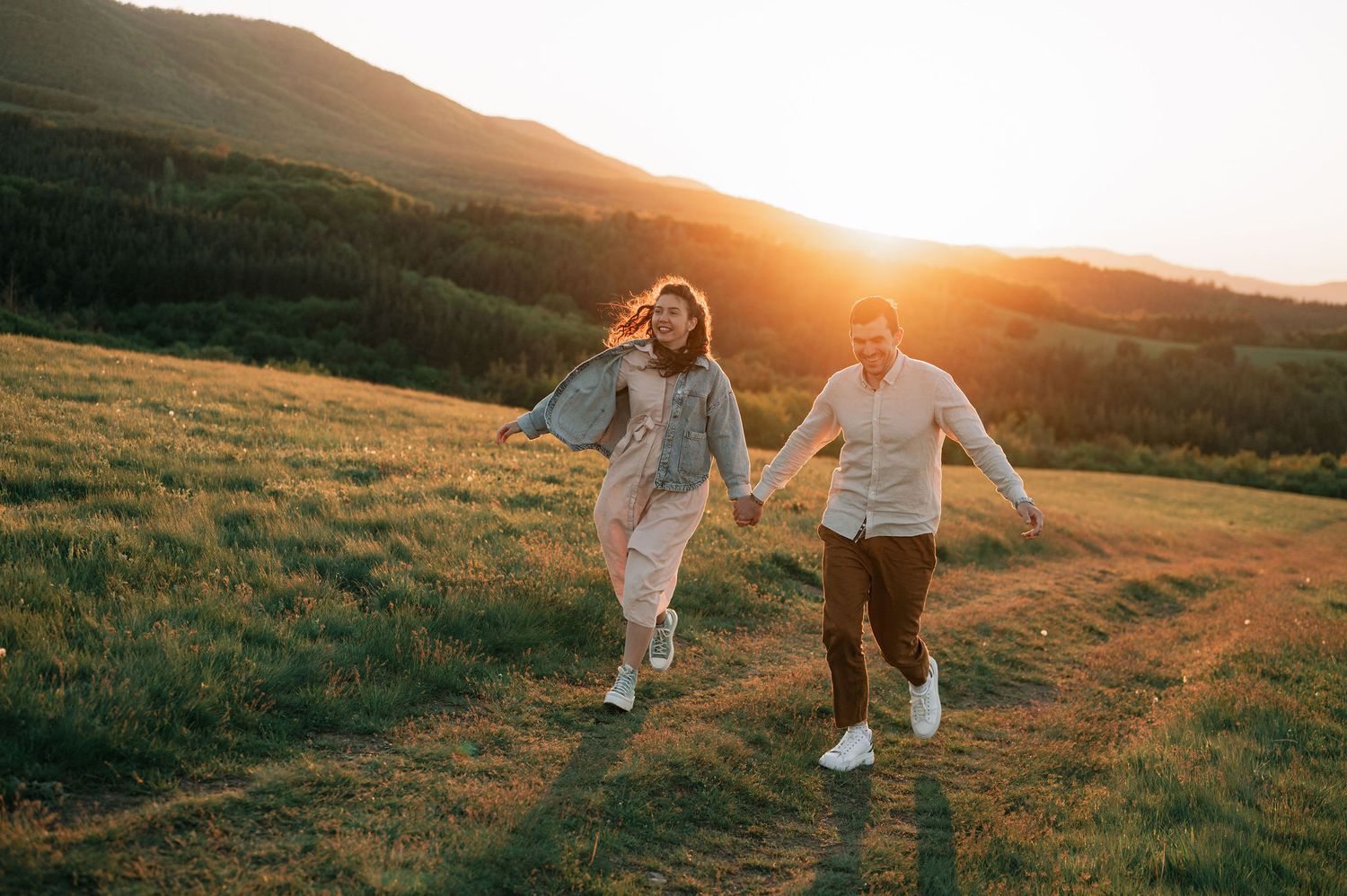 Couple running playfully through a grassy hillside meadow at golden sunset.
