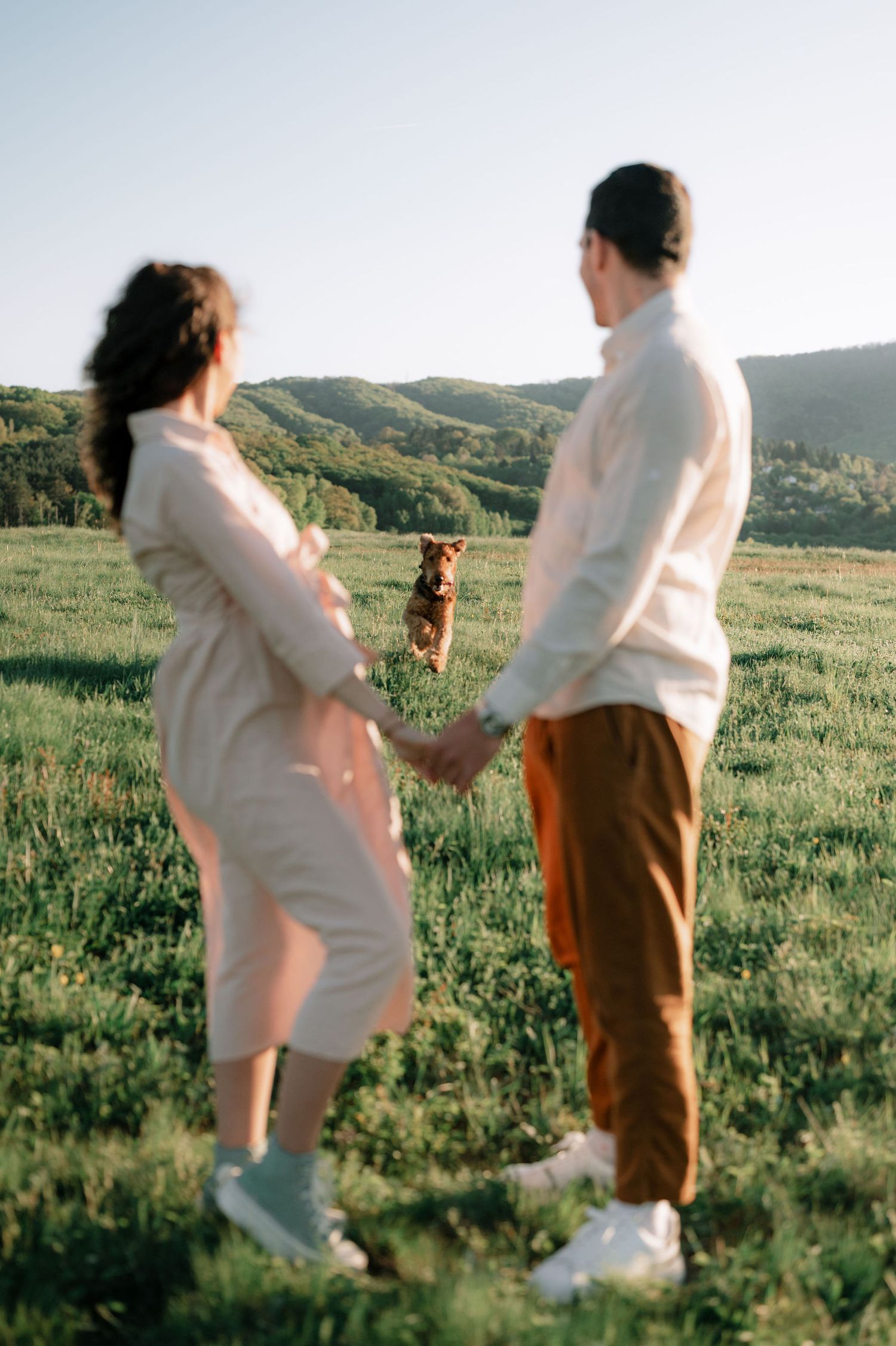 A couple in light colored clothing stands together in a grassy meadow during golden hour with mountains in the background.