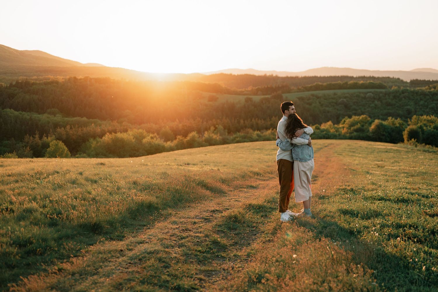 A couple walks along a scenic mountain trail at sunset with golden light illuminating the rolling hills and landscape.