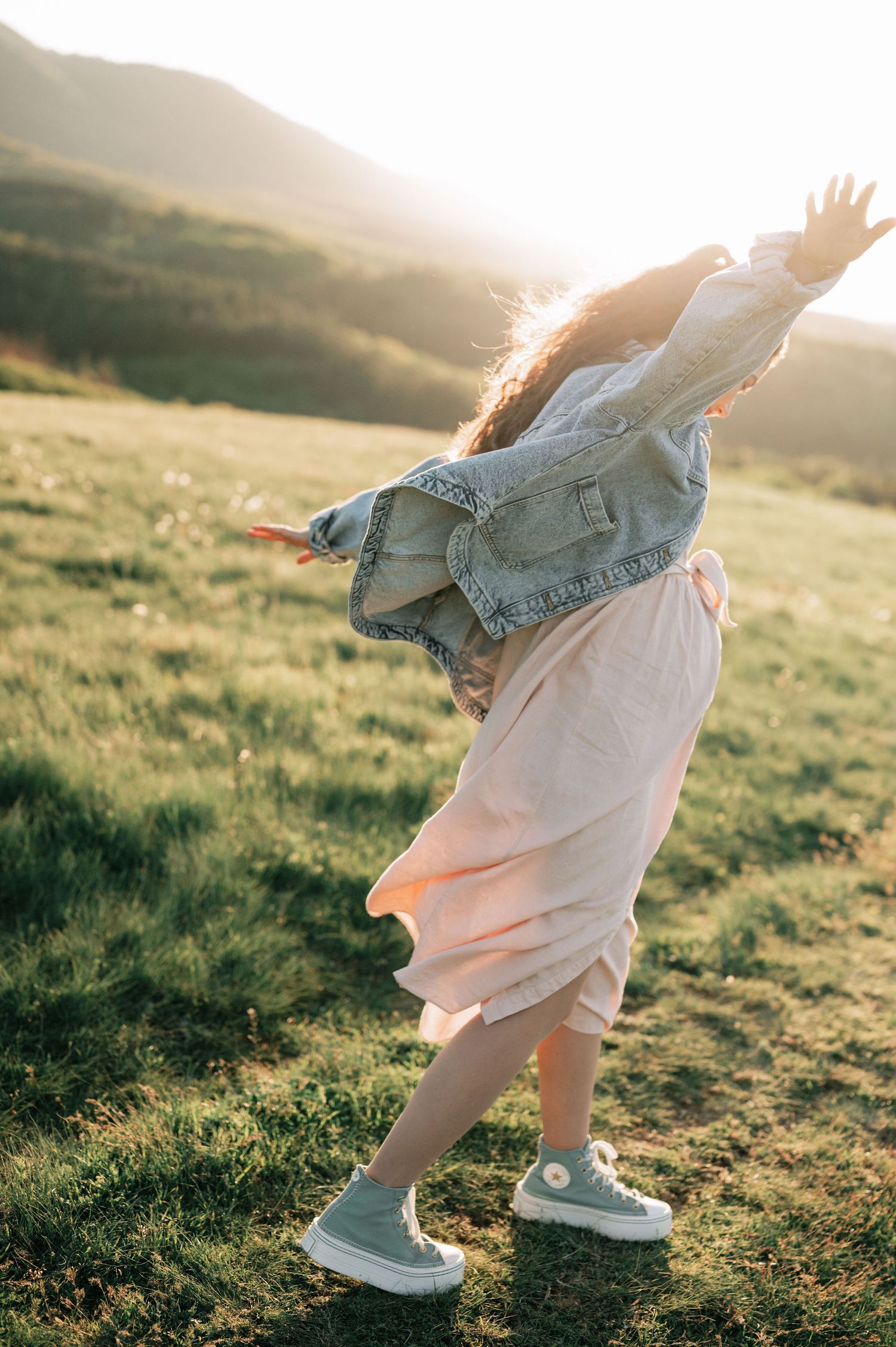 Person in denim jacket and white dress twirls joyfully in a sunlit meadow at golden hour.
