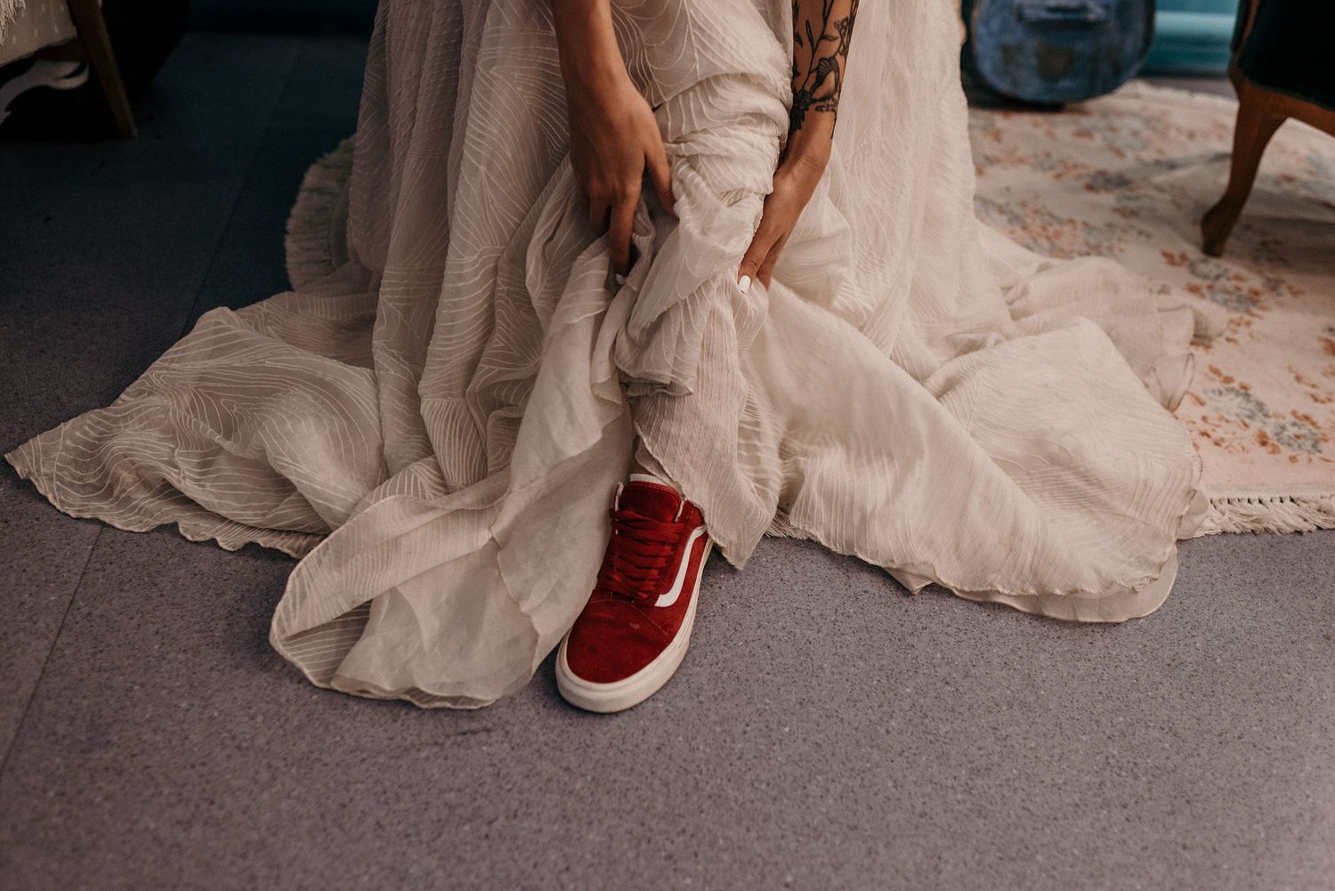 Red sneakers peek out from under flowing white wedding dress on grey carpet.