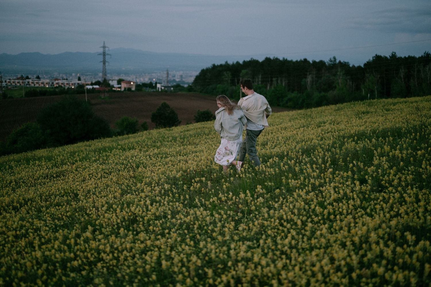 A couple enjoys a peaceful moment in a sprawling field of yellow flowers with mountains visible.