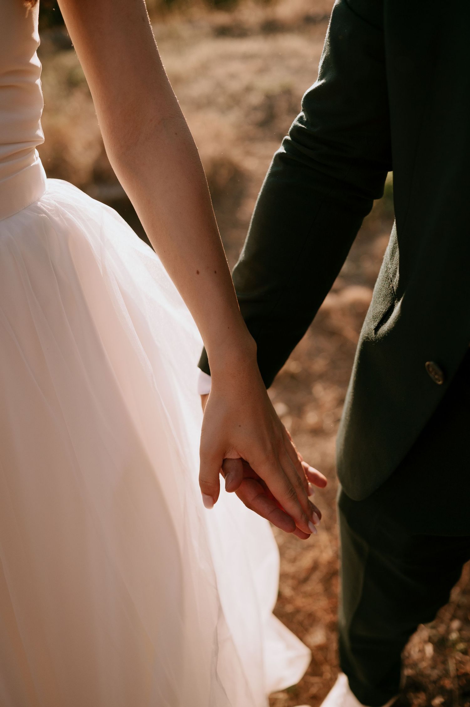 Close-up of bride and groom holding hands, showing white dress and dark suit details.