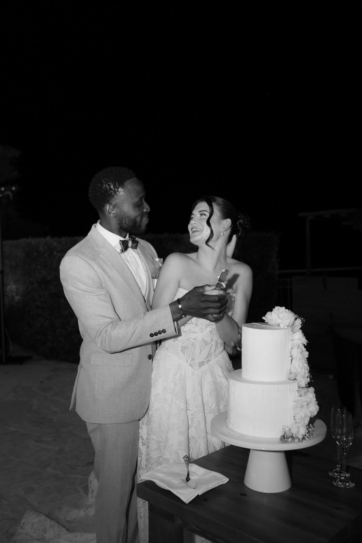 Black and white photo of a bride and groom smiling together beside a tiered wedding cake at their outdoor reception.