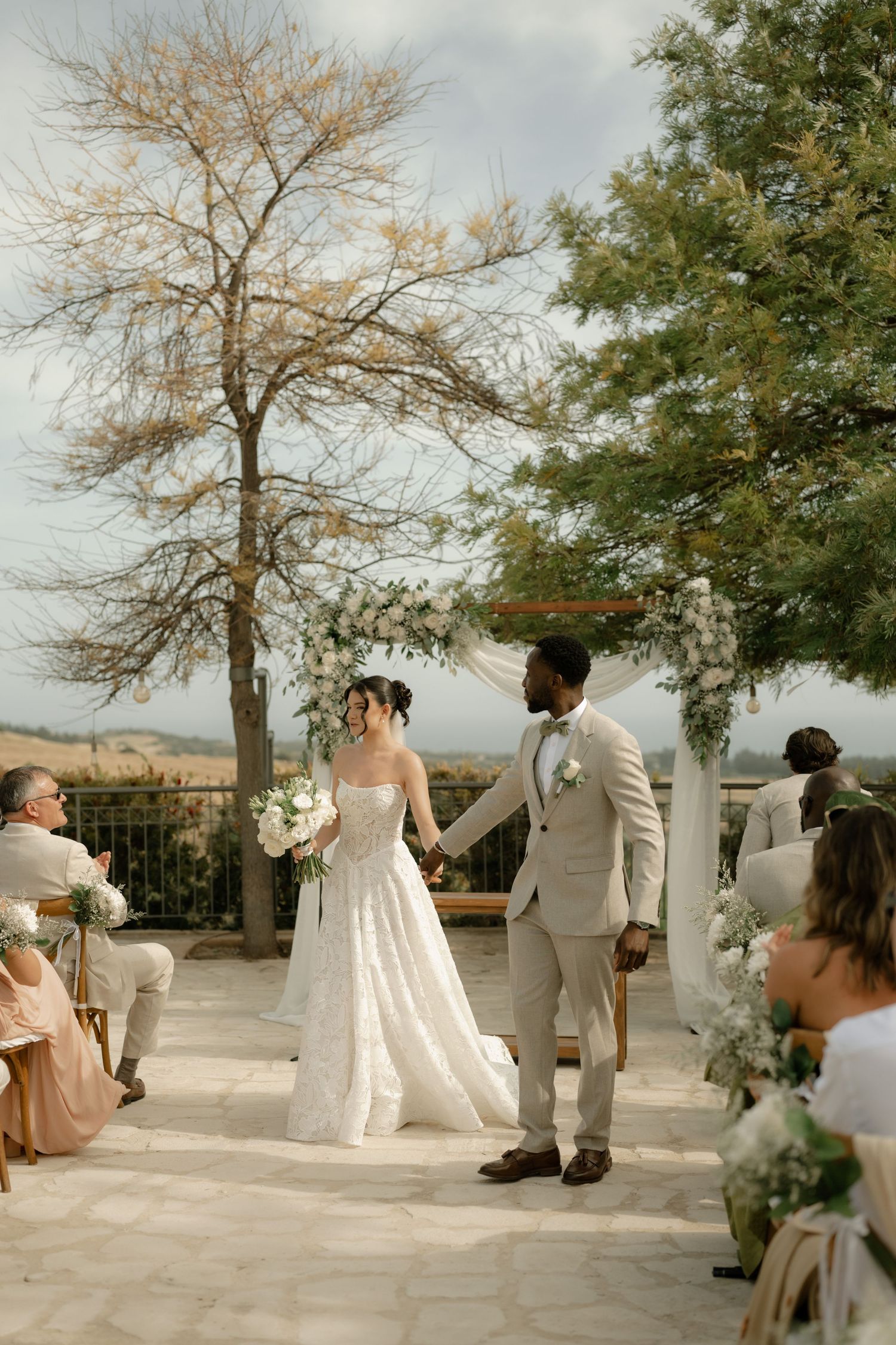 Bride and groom hold hands at outdoor wedding ceremony with floral arch, white blooms, and scenic landscape backdrop.