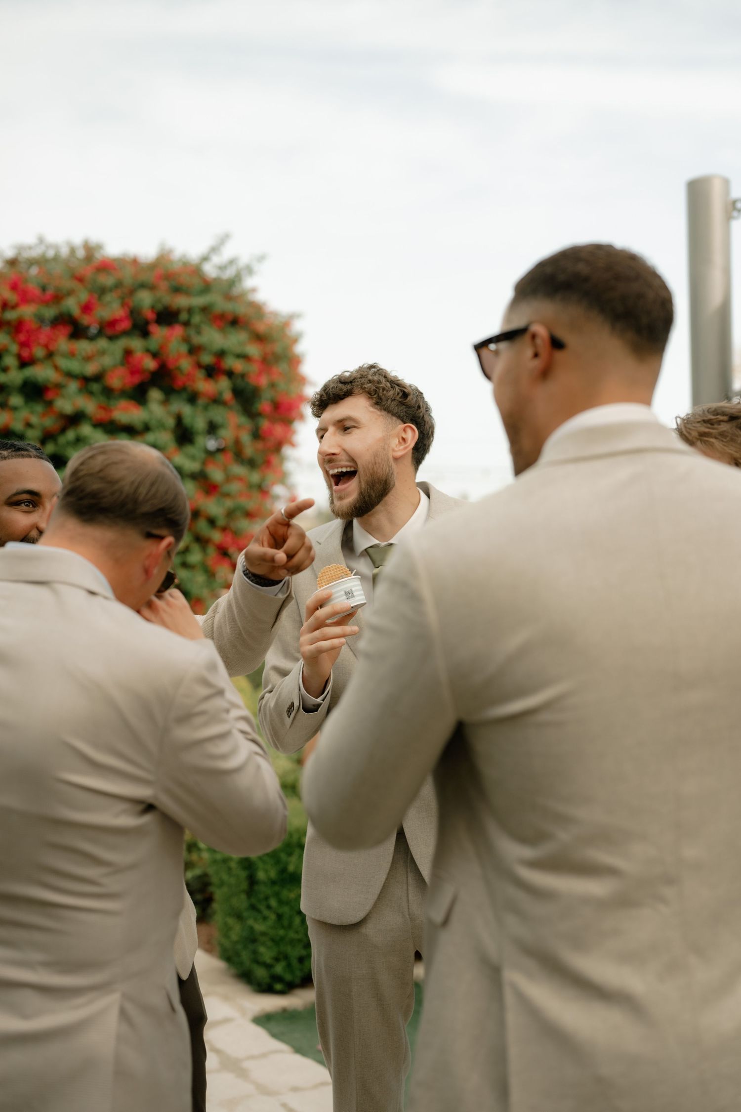 Groomsmen in tan suits laughing and celebrating outdoors with red floral backdrop at a wedding.