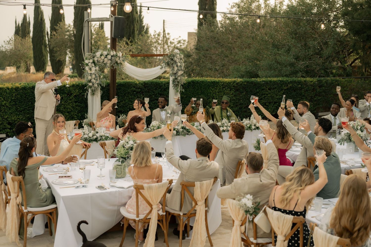 Outdoor wedding reception toast with guests raising glasses at elegantly decorated tables surrounded by lush greenery.