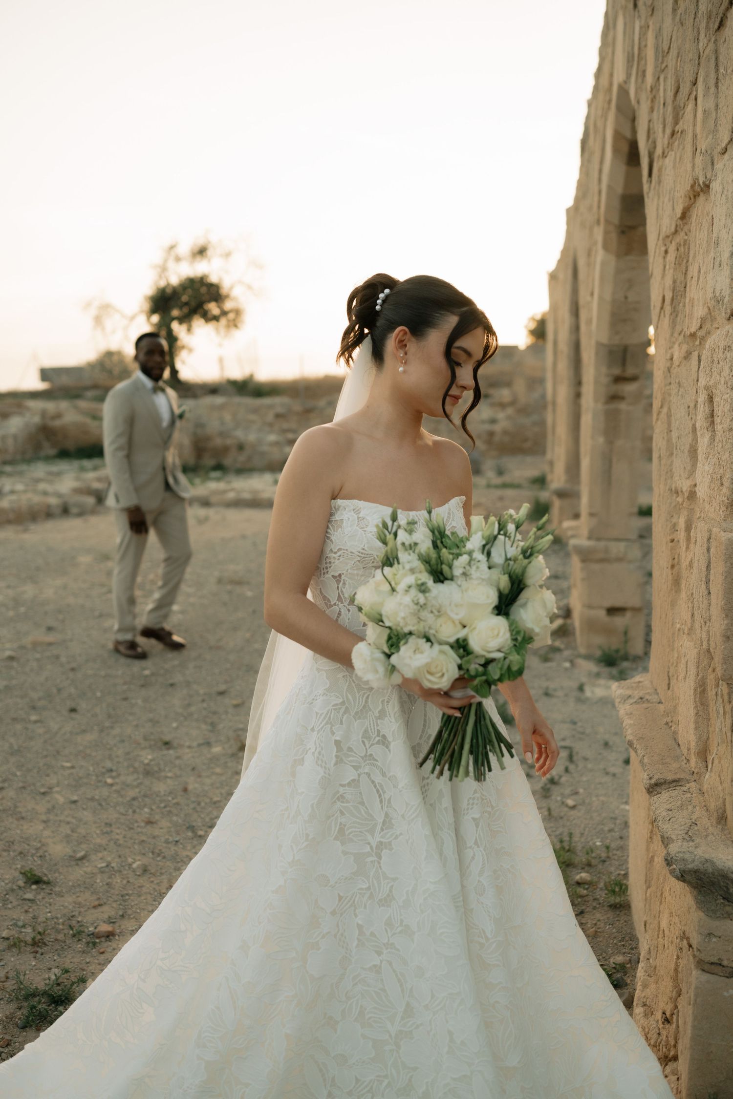Bride in strapless lace wedding gown holds white bouquet at ancient ruins, groom in tan suit stands in background.
