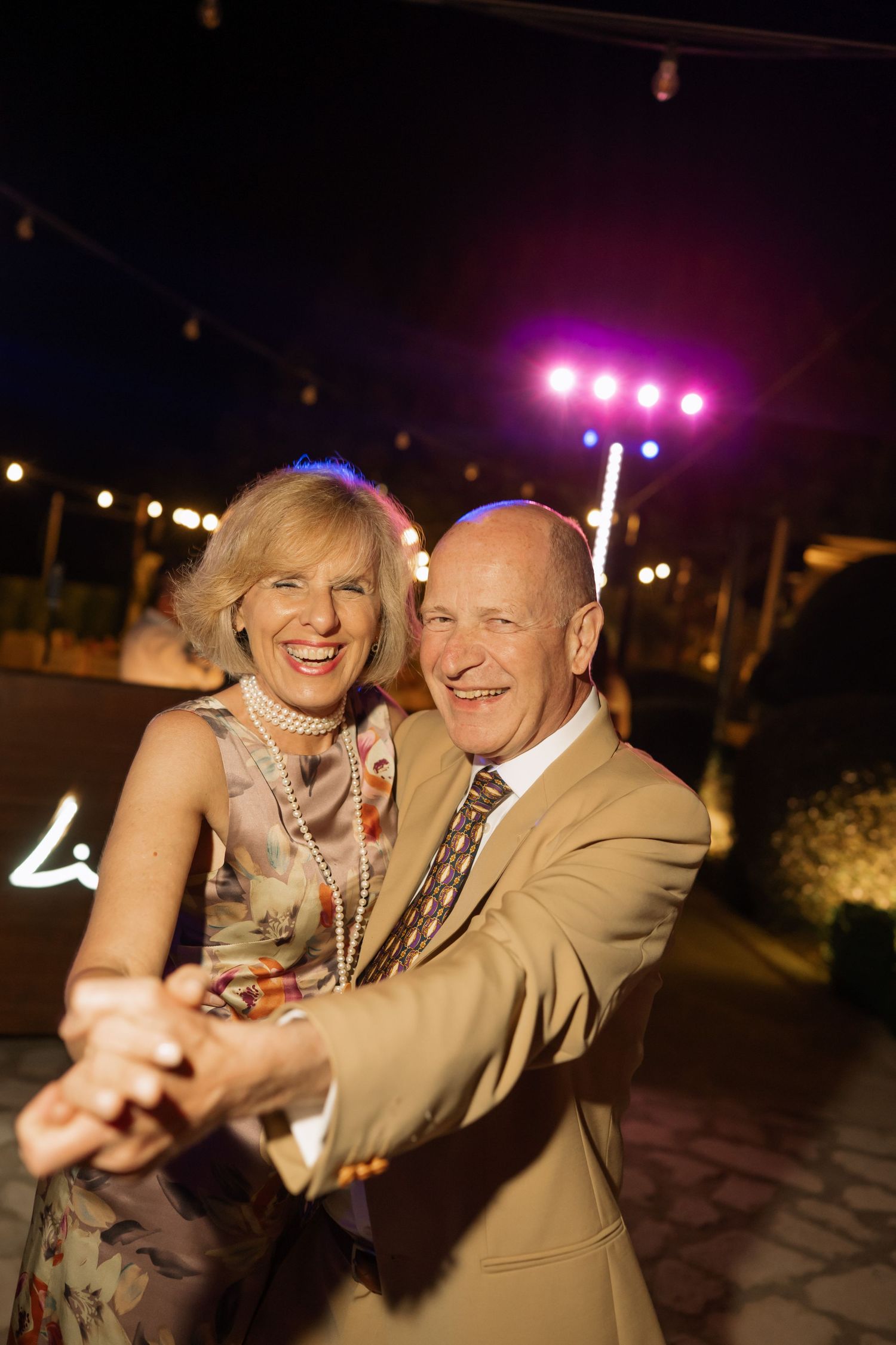 Happy senior couple dancing together outdoors at a night event, smiling under colorful party lights.