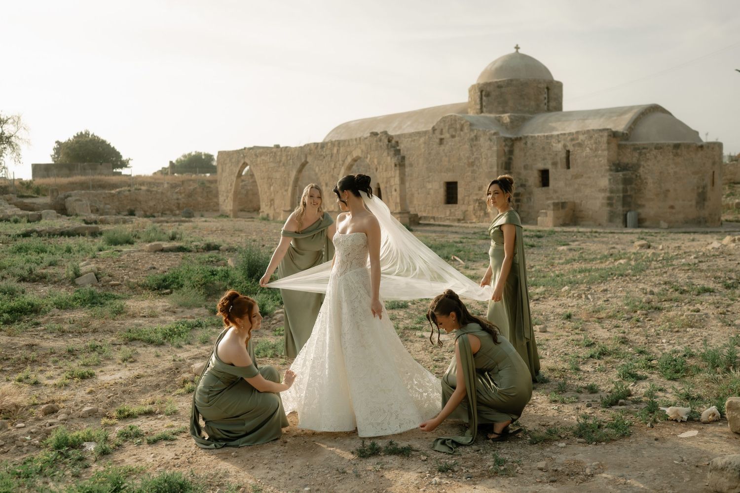 Bride in white gown with bridesmaids in sage green adjusting veil near ancient stone church ruins in desert landscape.