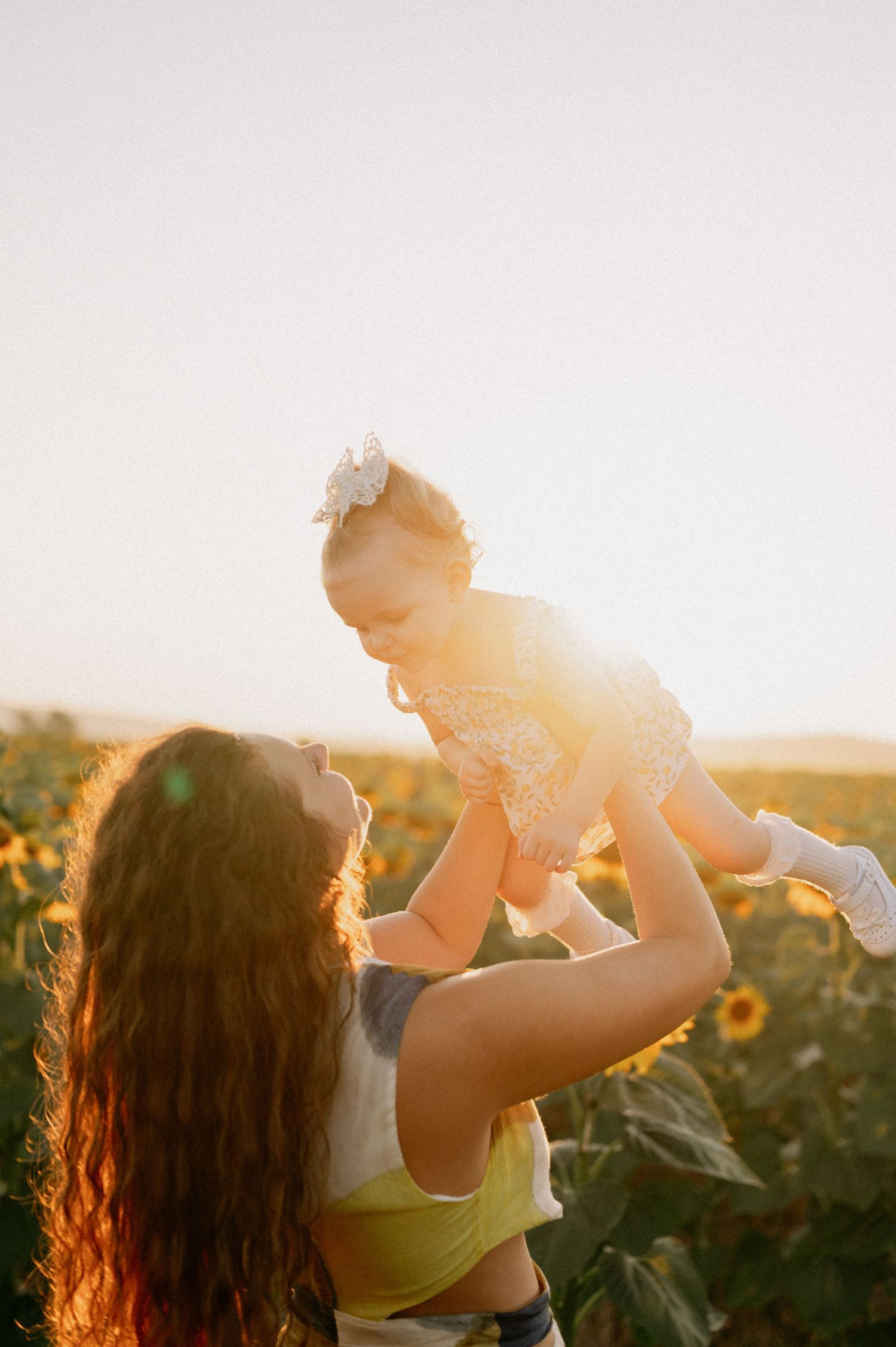 A mother lifts her baby girl in a sunflower field, bathed in golden sunset light.