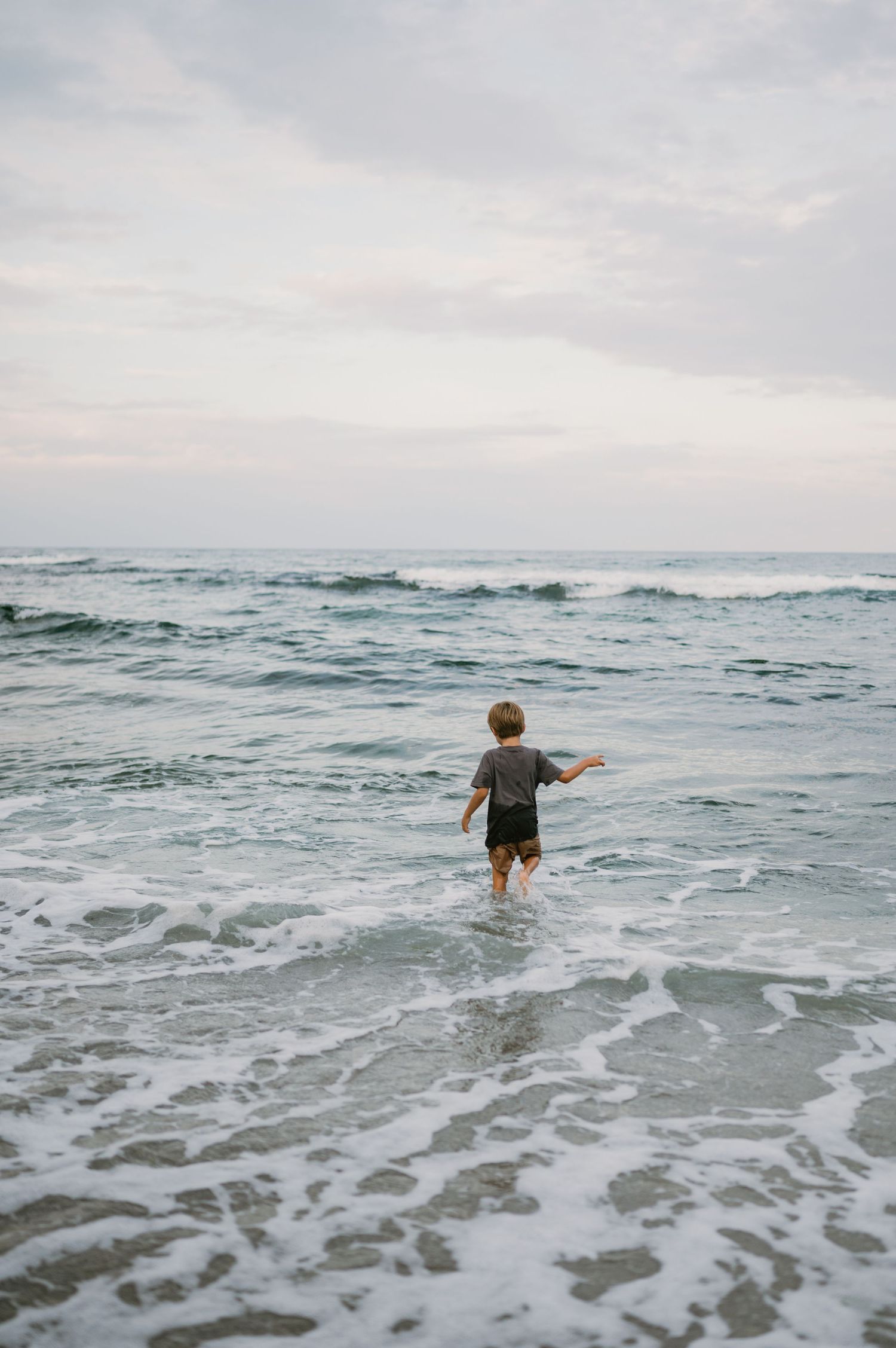 Young boy wading into ocean waves at the beach during overcast weather, viewed from behind.