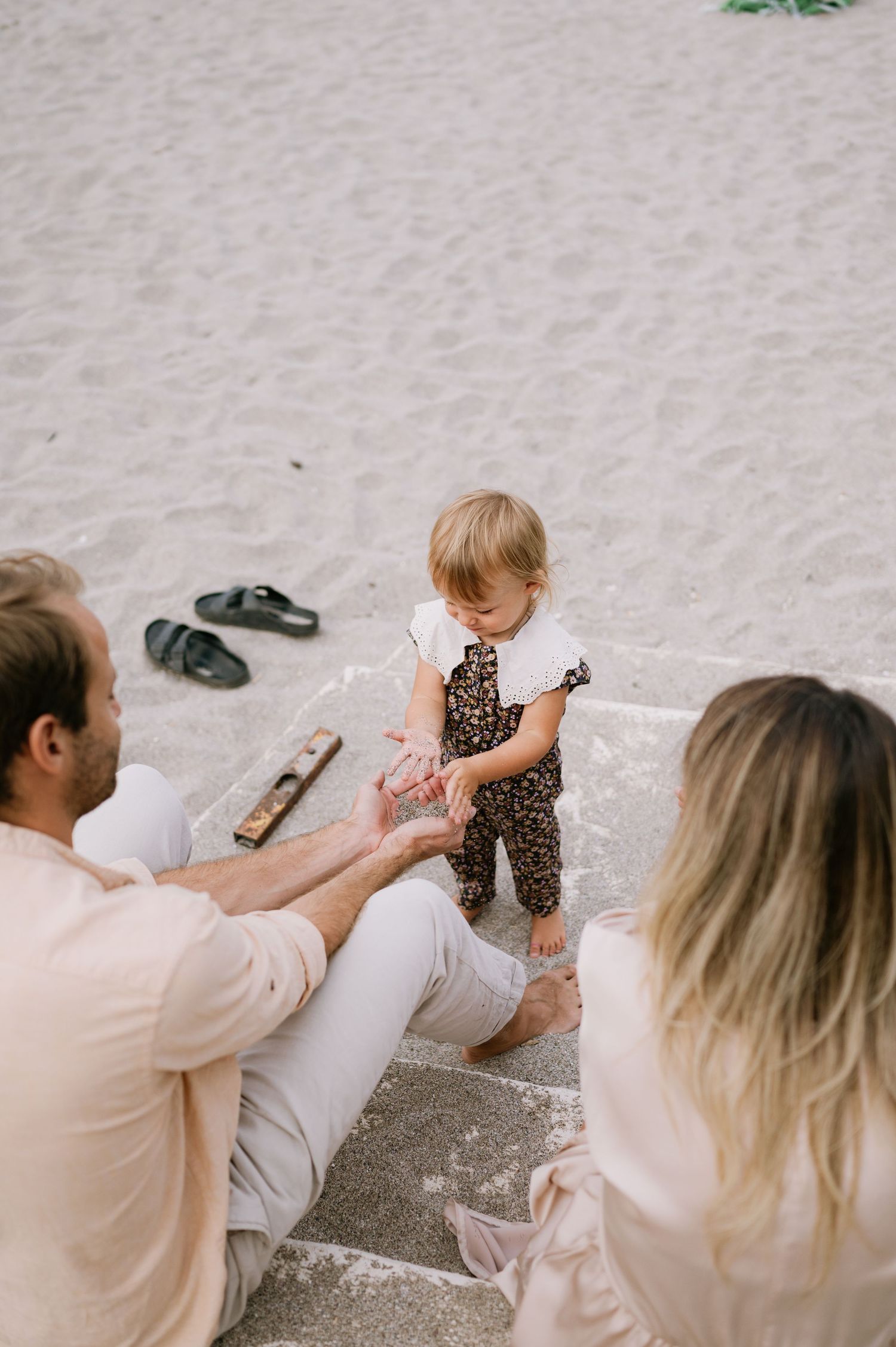 A toddler in leopard print overalls stands on beach steps with parents sitting nearby on a sandy beach.