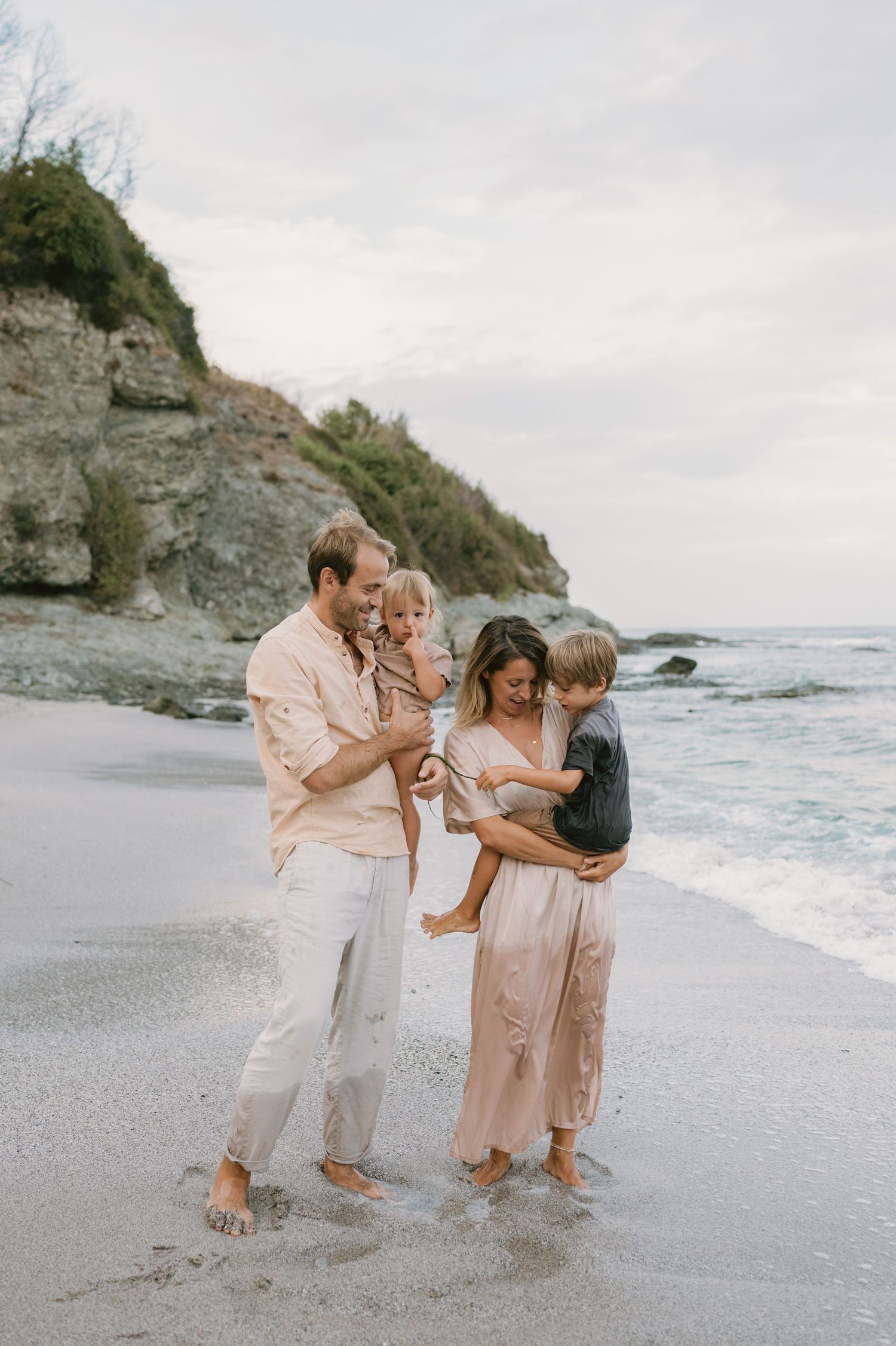 Family of four enjoying a beach day, parents holding young children near ocean waves with rocky cliffs in background.
