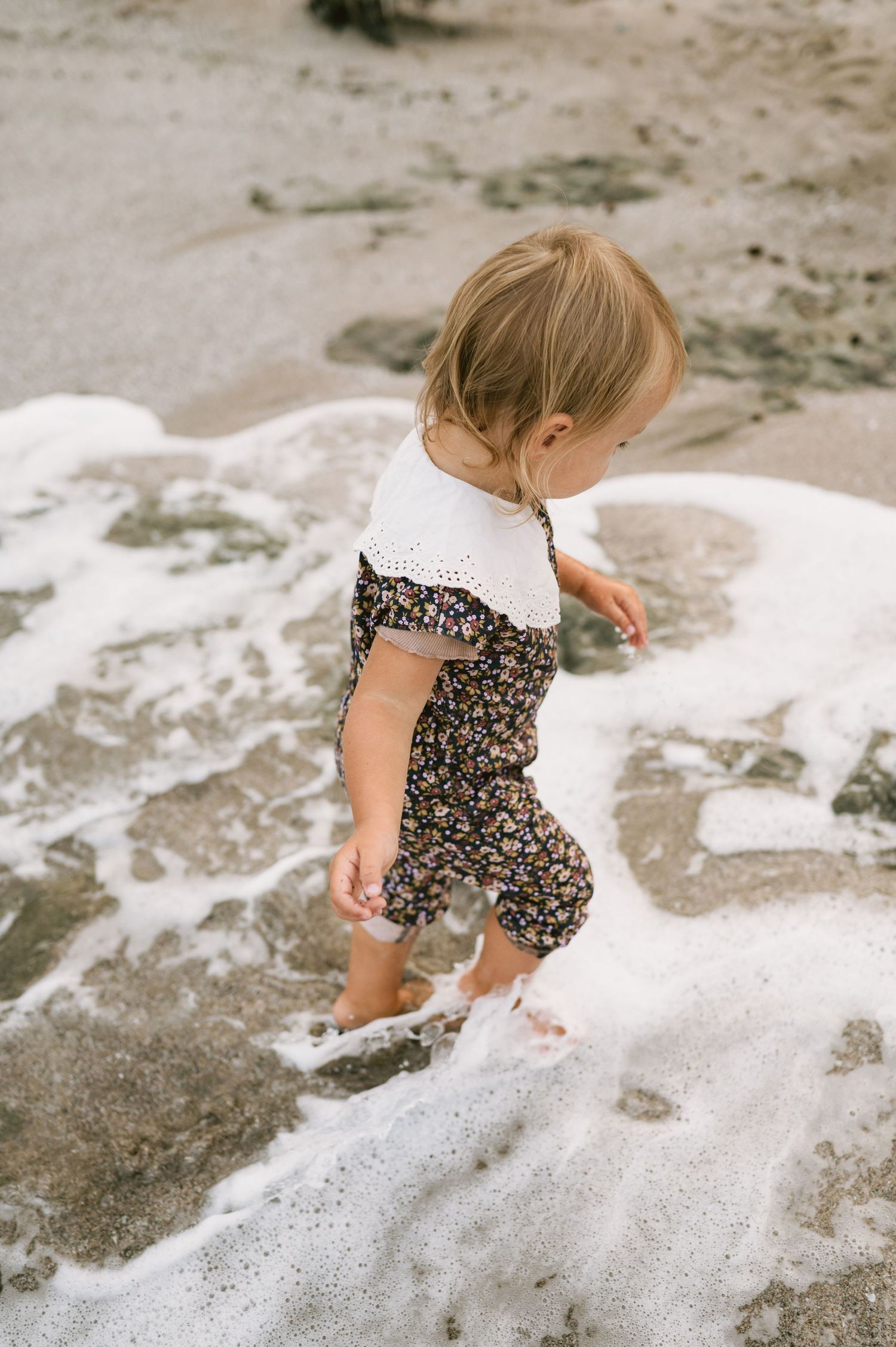 Toddler girl in floral romper playing in ocean waves on a sandy beach.