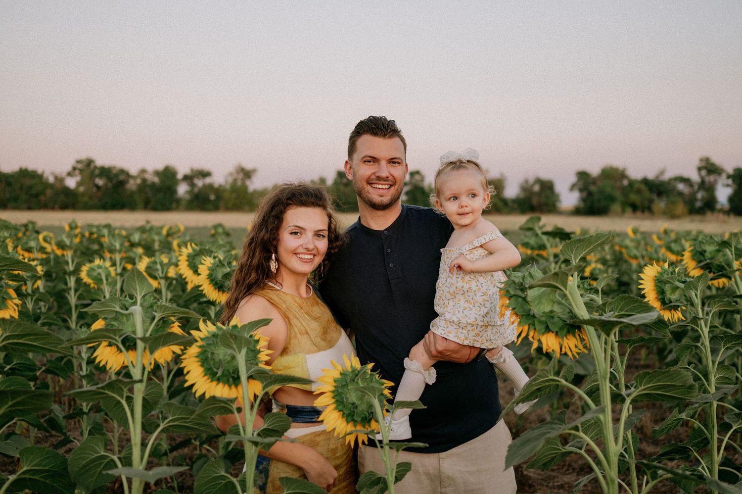 Family of three posing in a sunflower field at sunset, parents smiling with baby girl in floral outfit.
