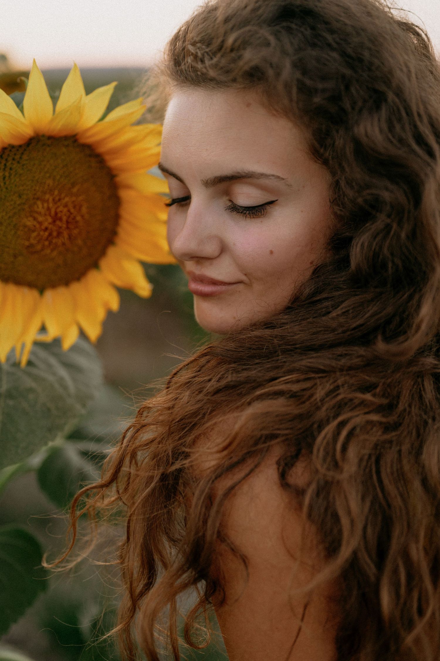 Young woman with curly hair smiling serenely beside a bright yellow sunflower in a field.