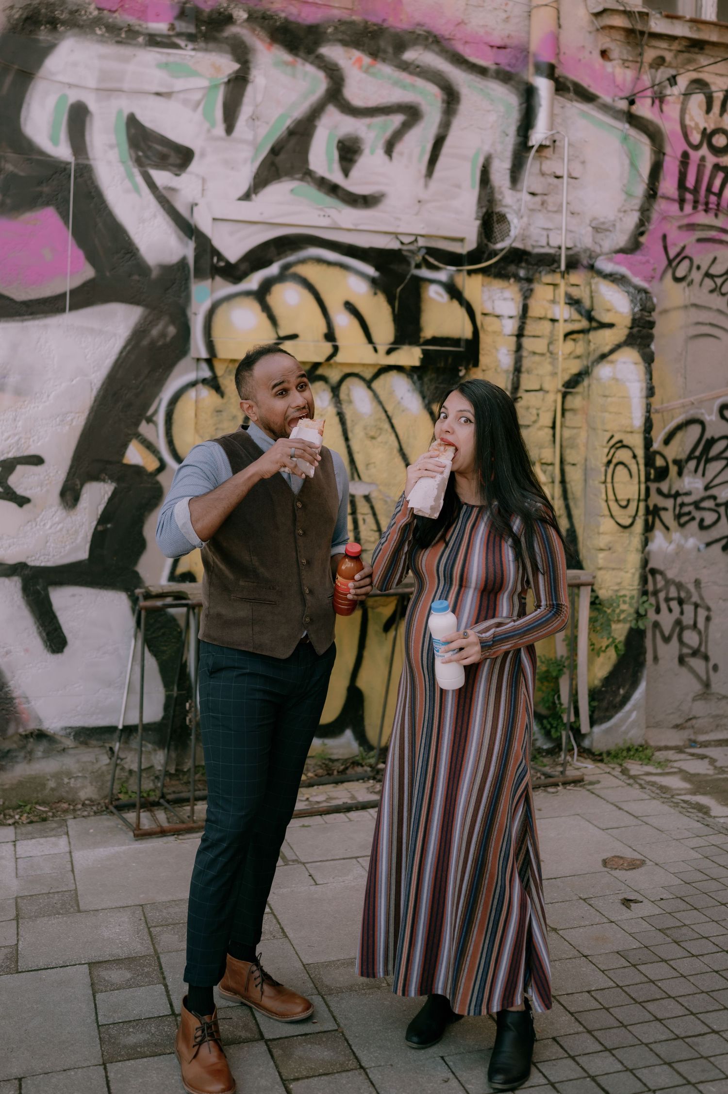 A couple eating street food in front of a colorful graffiti wall, woman in striped dress, man in vest and trousers.