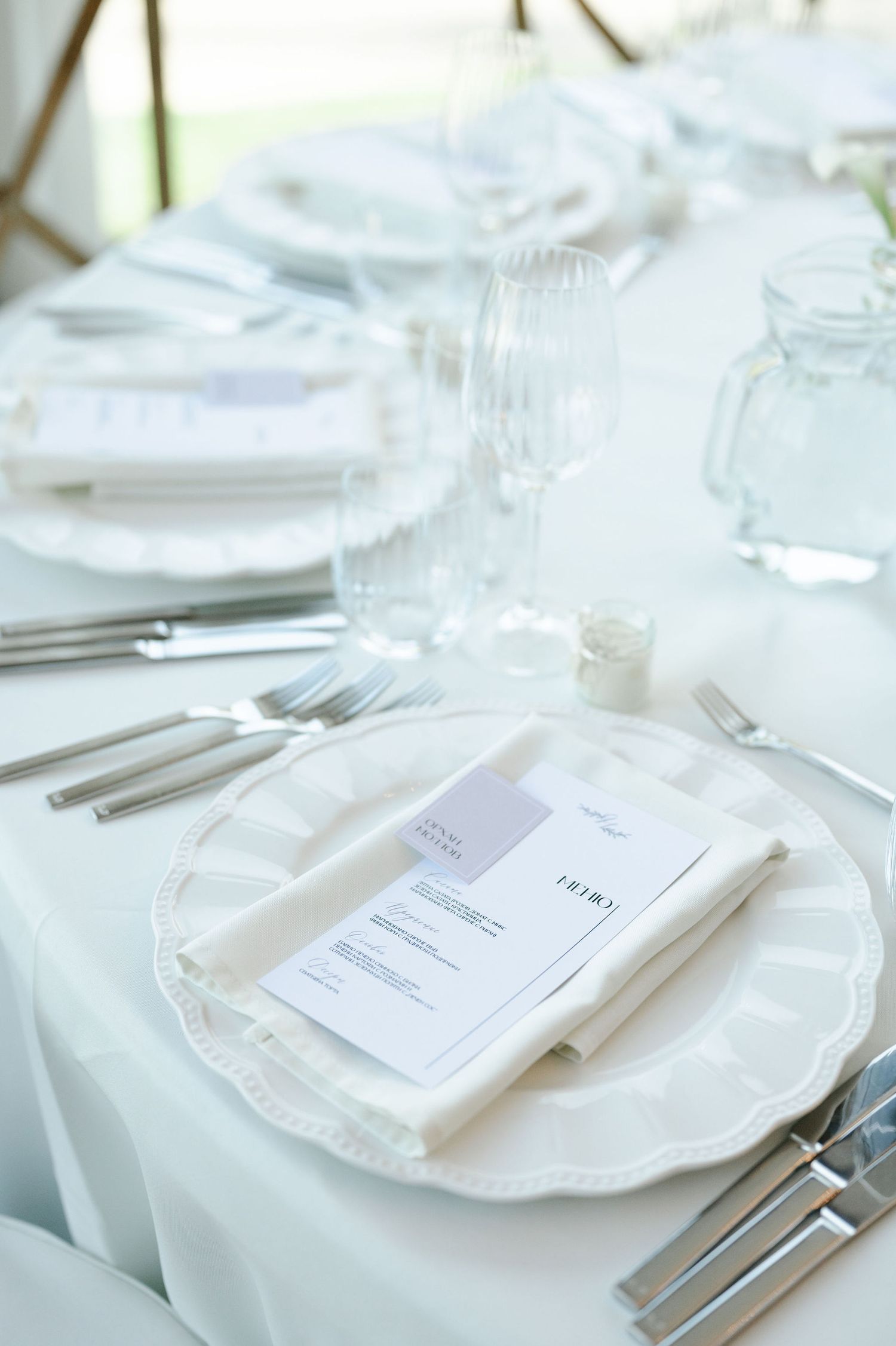 Close-up of minimalist white place settings with menu cards and silverware on scalloped plates.
