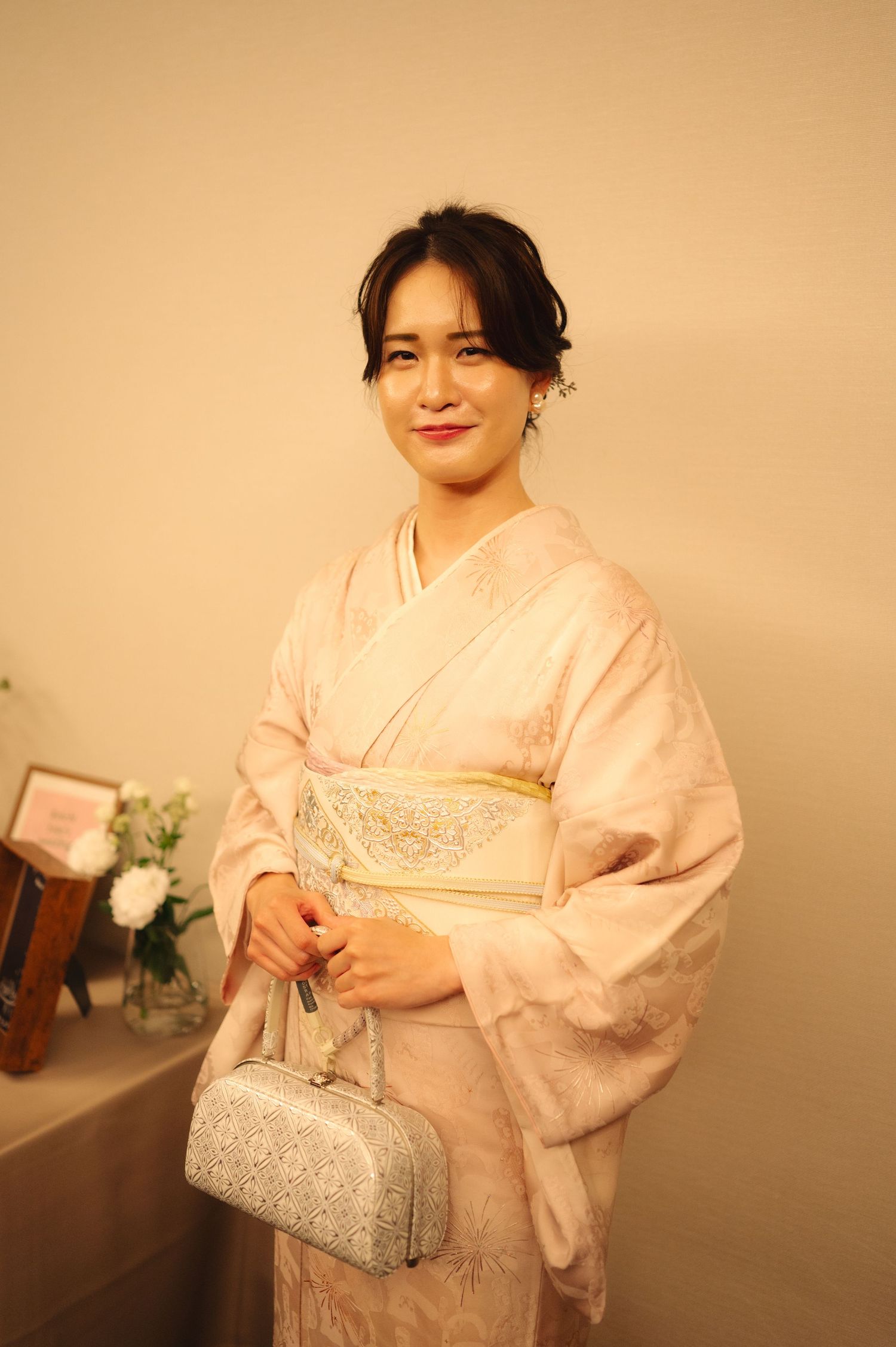 Person wearing a pale pink kimono and silver clutch bag poses against beige wall.